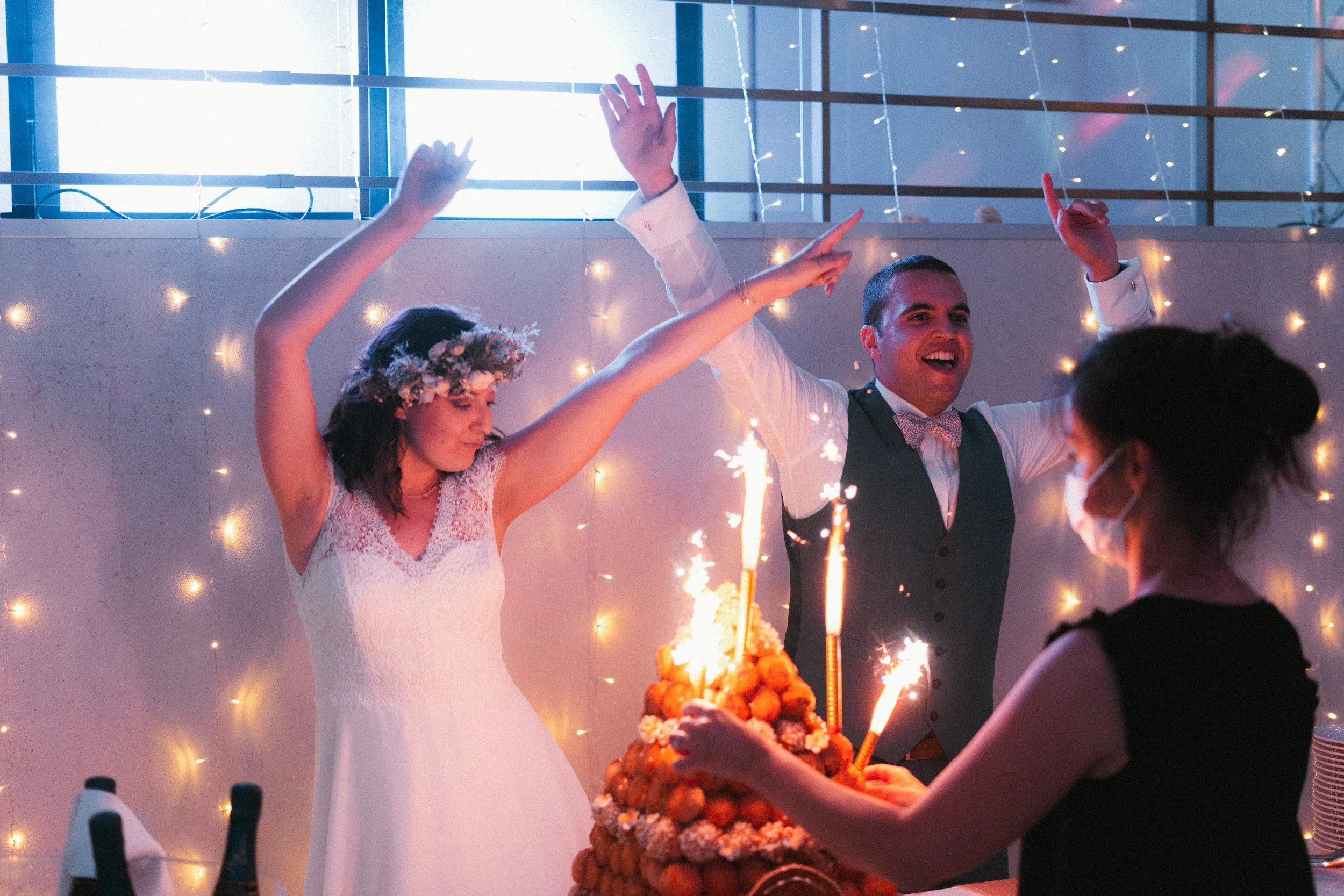 Un couple de mariés célèbre leur mariage avec un gâteau et des feux d'artifice, entourés d'invités, dans une salle décorée de lumières.