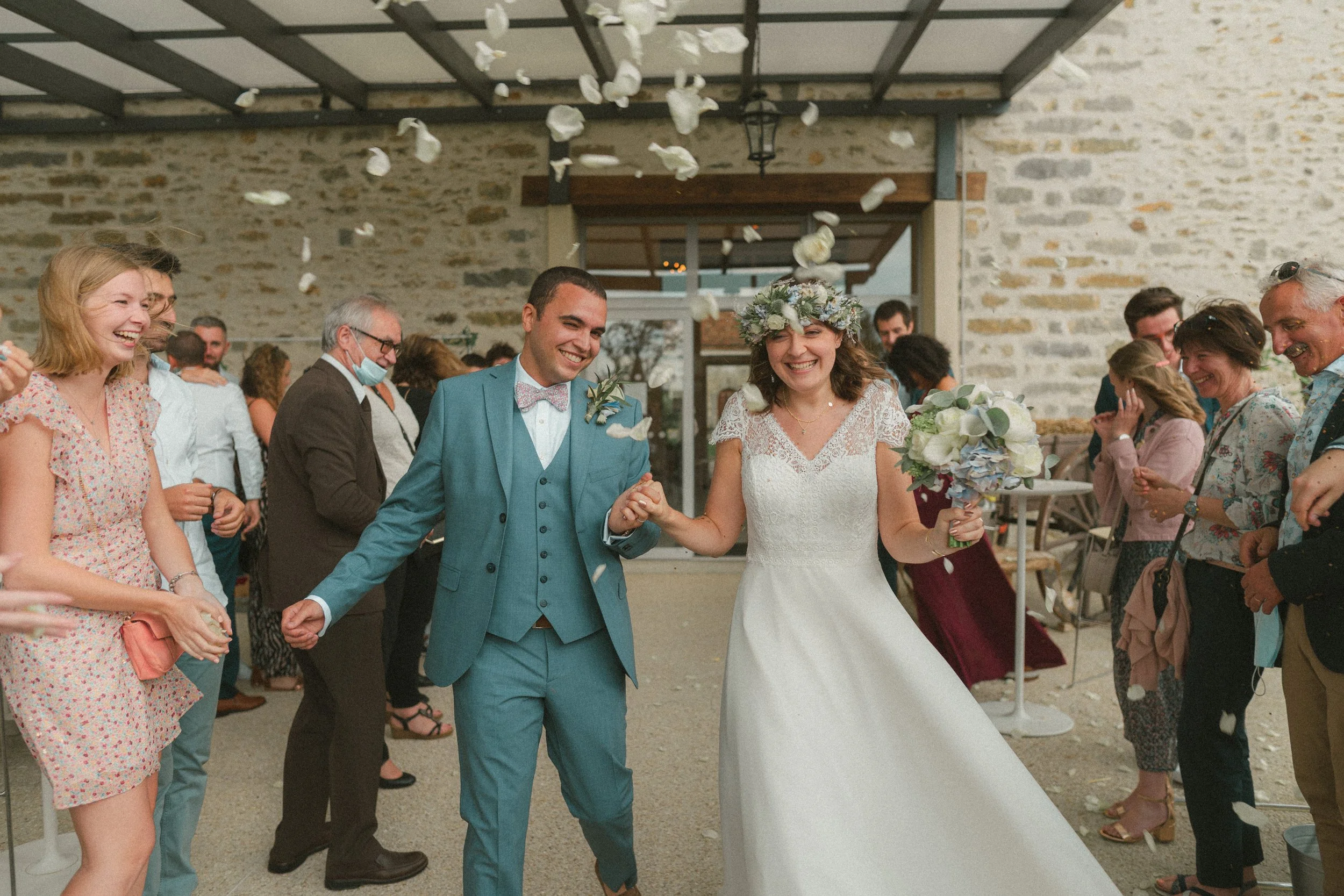 Un couple de mariés souriants marche main dans la main au milieu d'invités lors d'un mariage en intérieur, décor avec des pétales de fleurs tombant du plafond.