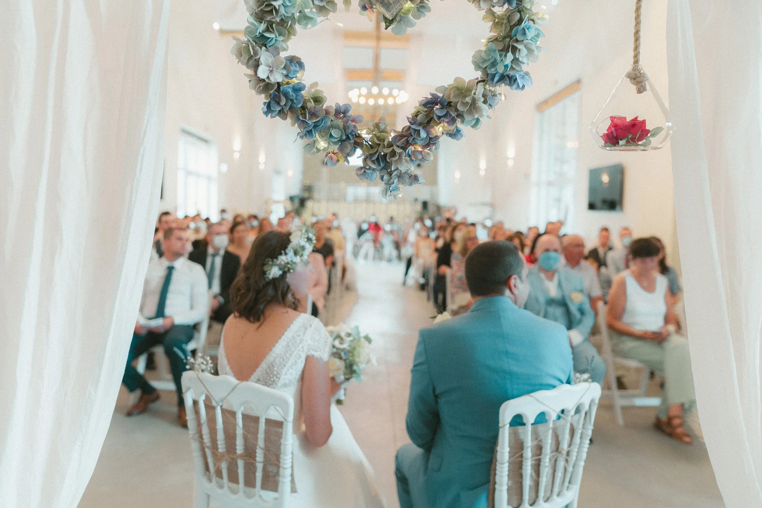 Un mariage avec la mariée et le marié assis face à face sous une arche décorée de fleurs, dans une grande salle lumineuse remplie d'invités. La décoration inclut un cœur en fleurs suspendu au plafond et un petit vase avec une rose.