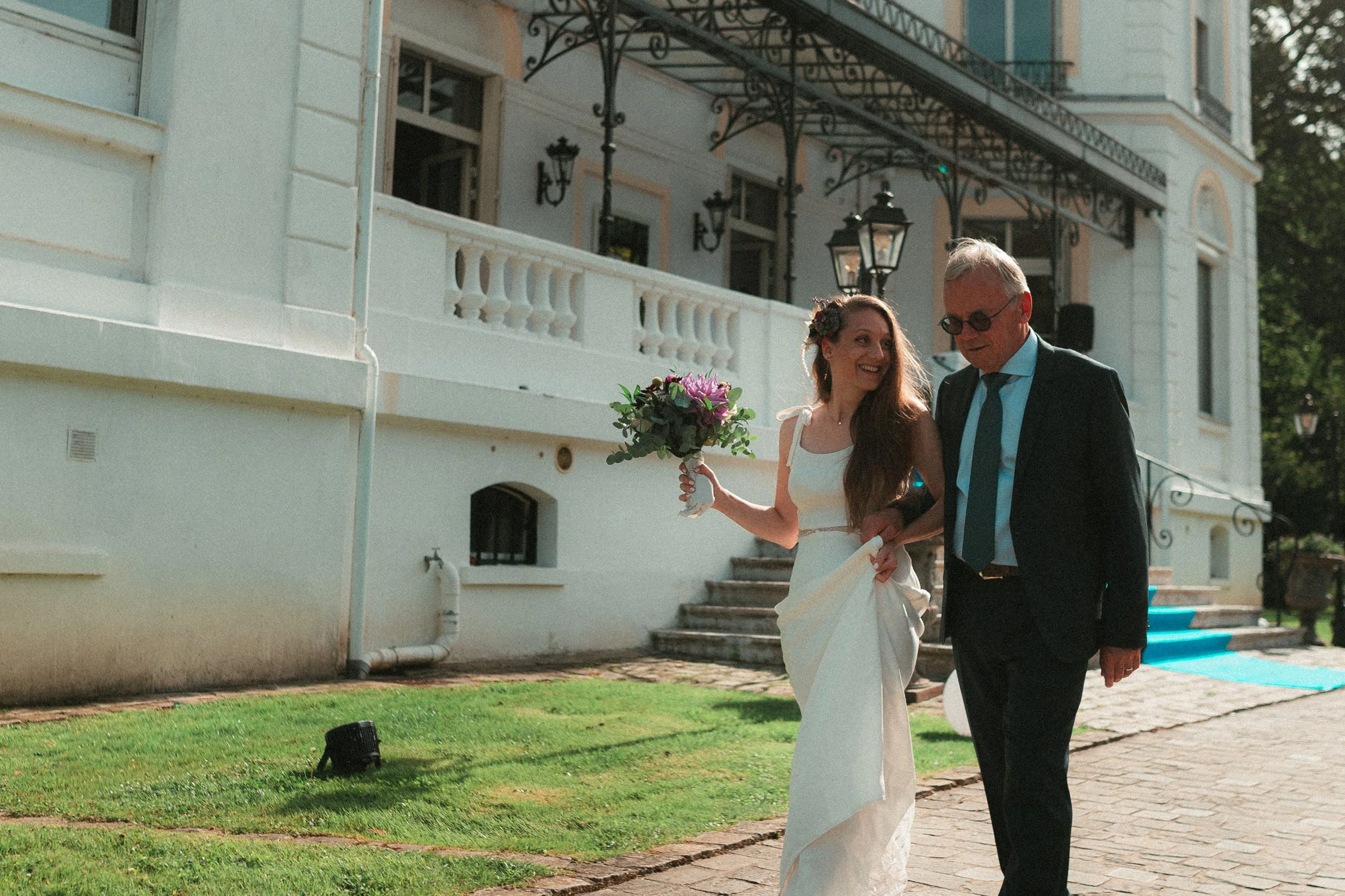 Une mariée souriante en robe blanche tenant un bouquet de fleurs, étant accompagnée d'un homme en costume portant des lunettes, devant un bâtiment blanc élégant lors d'un mariage en plein air.