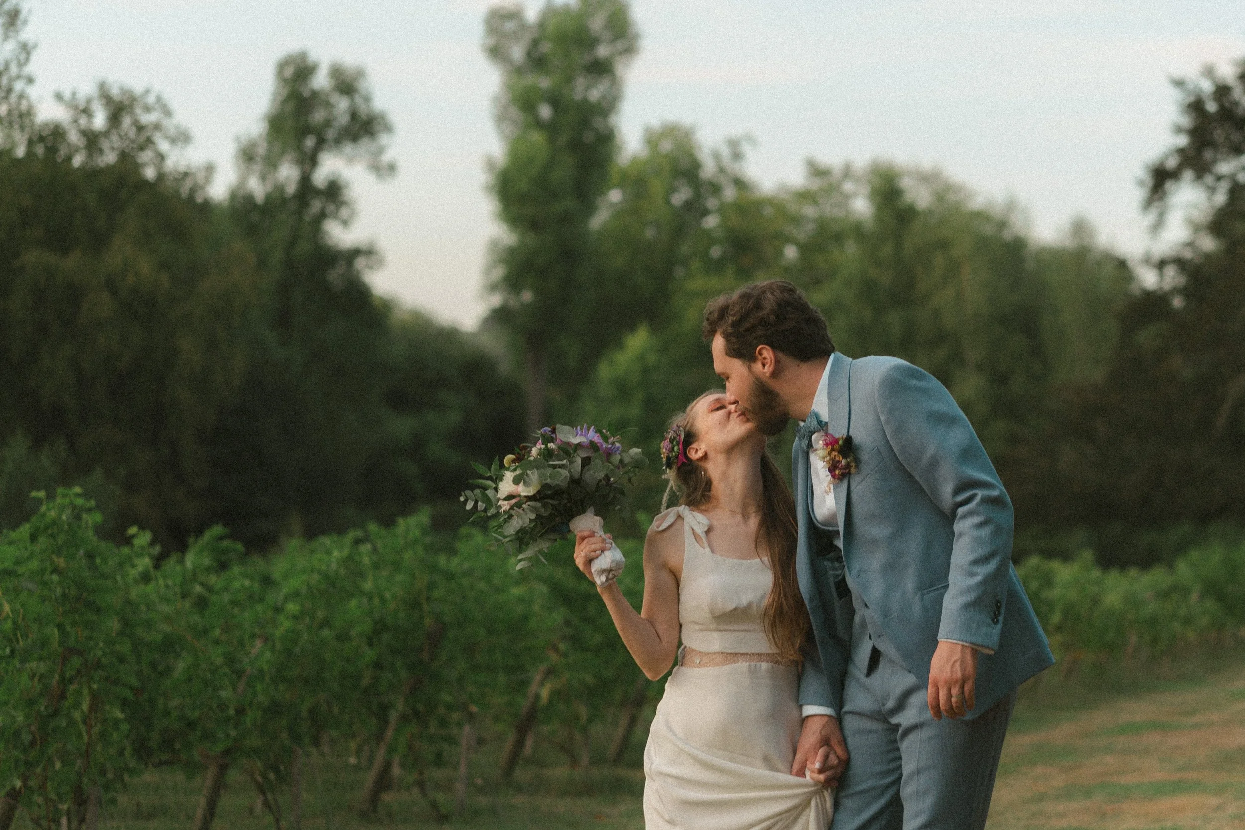 Un couple de mariés s'embrasse dans un champ en plein air, la femme tenant un bouquet de fleurs.