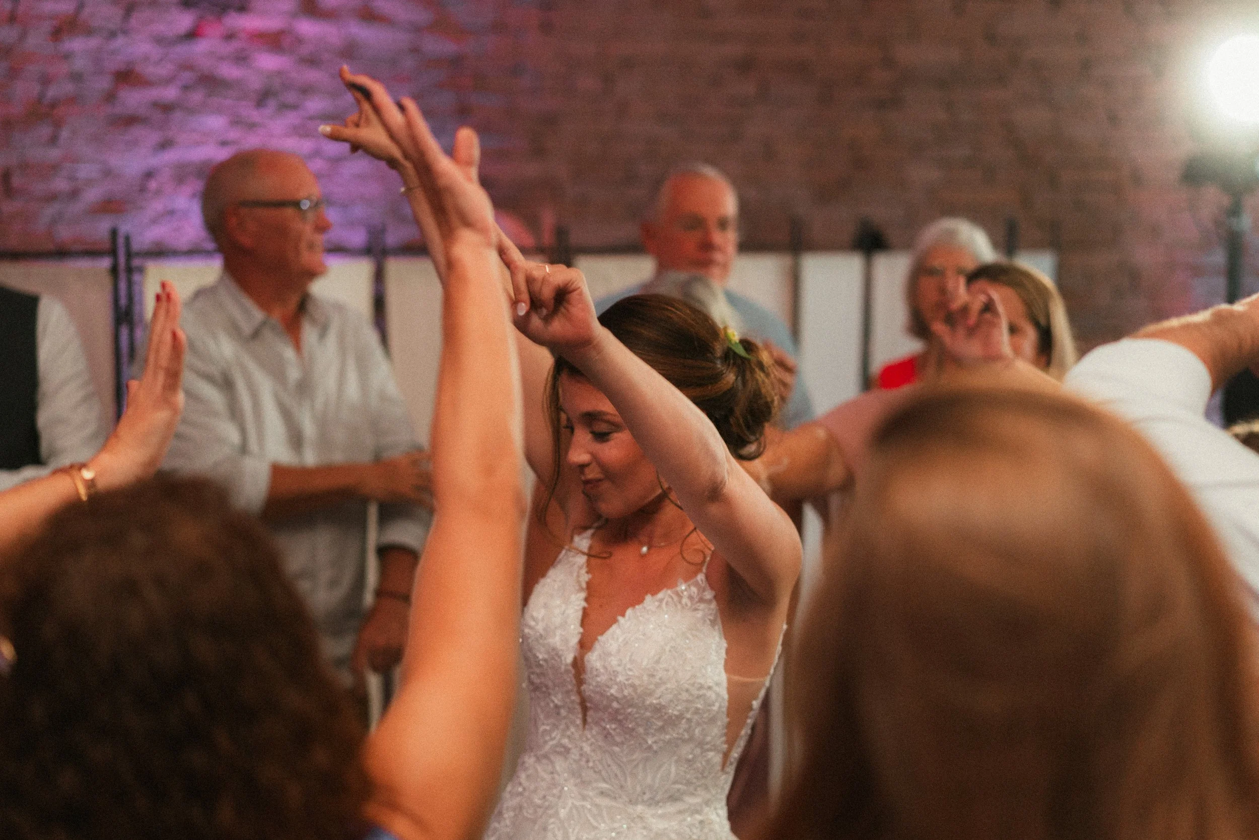 Une femme en robe de mariée danse lors d'une célébration, entourée de plusieurs personnes dans une pièce avec un mur en briques.