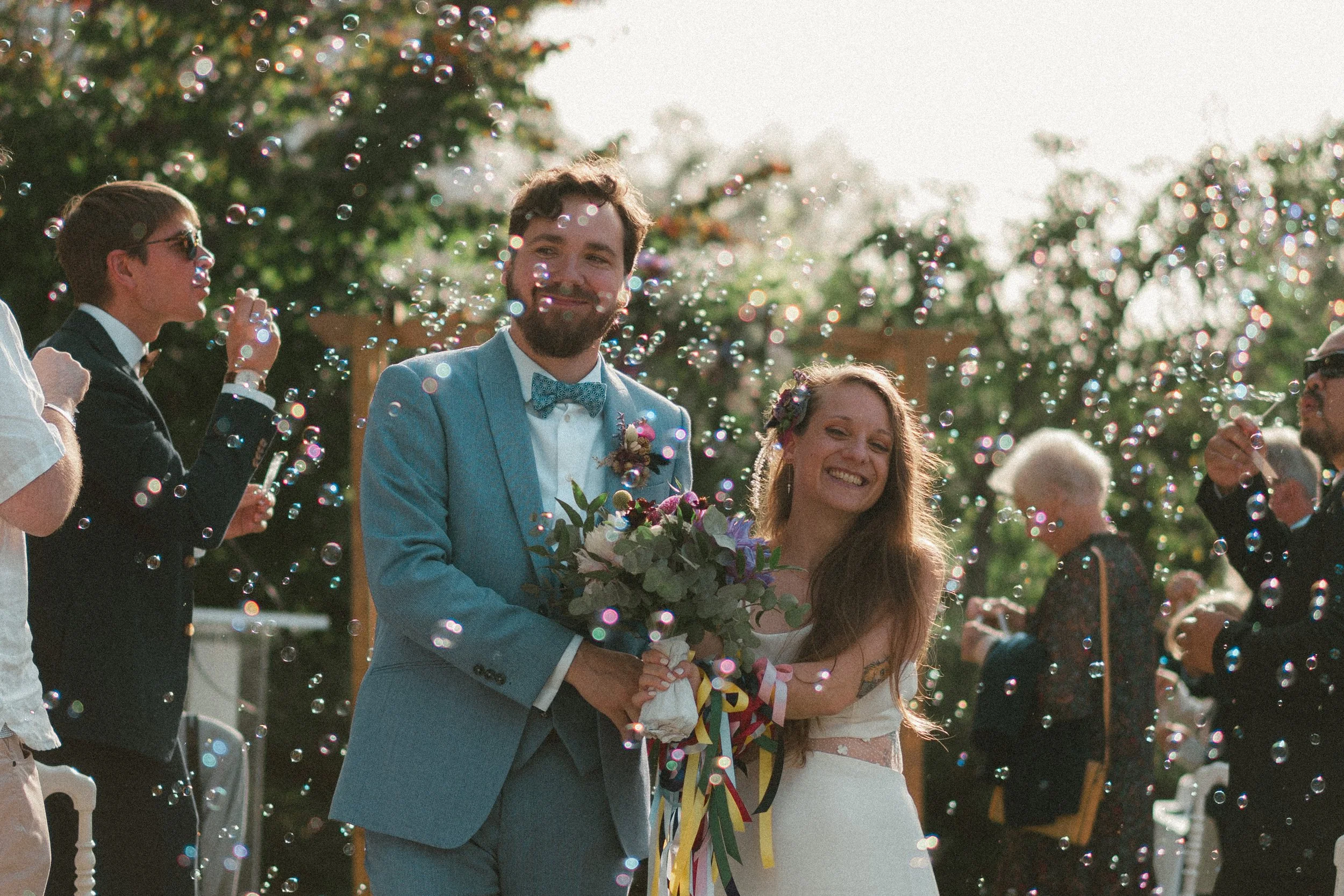 Un couple de mariés souriant, la femme tenant un bouquet de fleurs, entourés d'invités soufflant des bulles lors d'une célébration en plein air.
