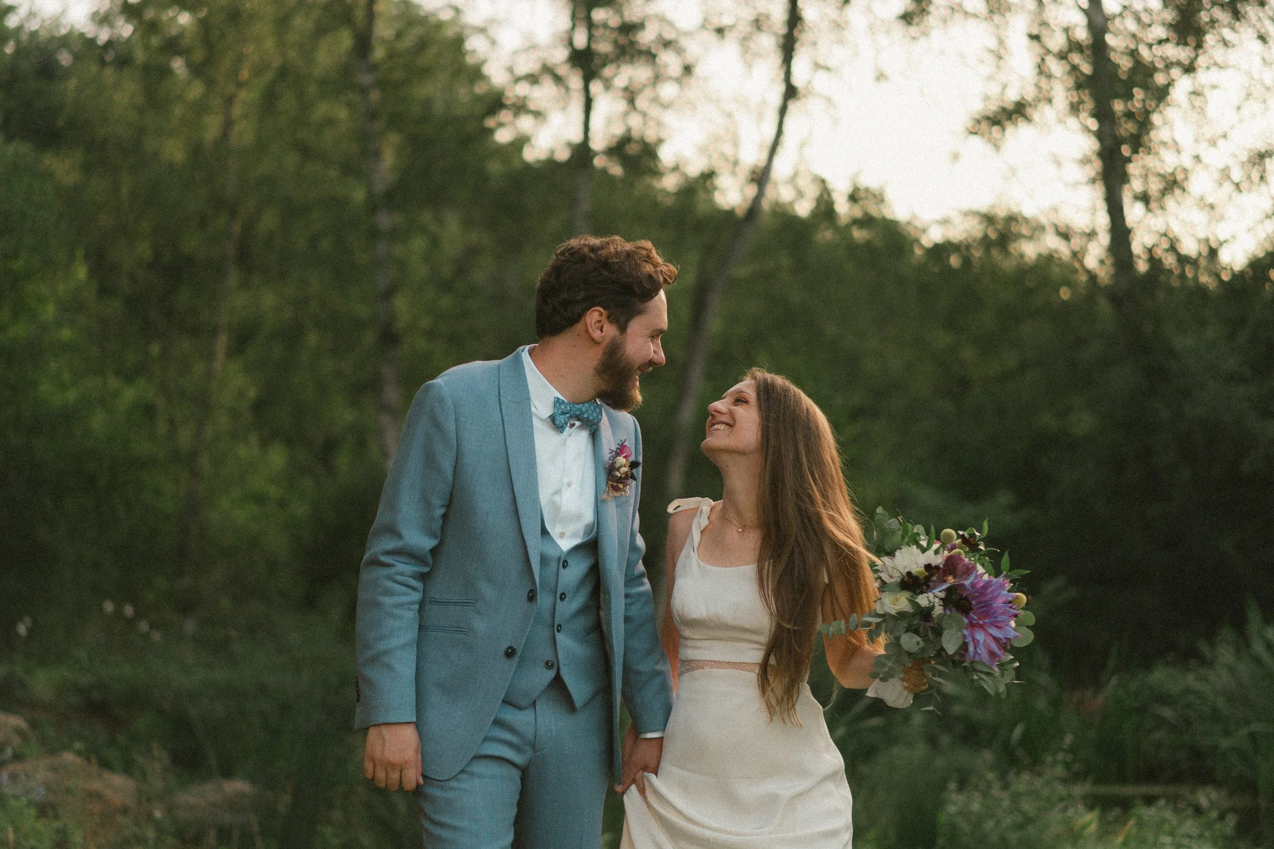 Un couple de mariés dans un endroit naturel en forêt lors de leur mariage, ils se regardent en souriant, la mariée tient un bouquet de fleurs.