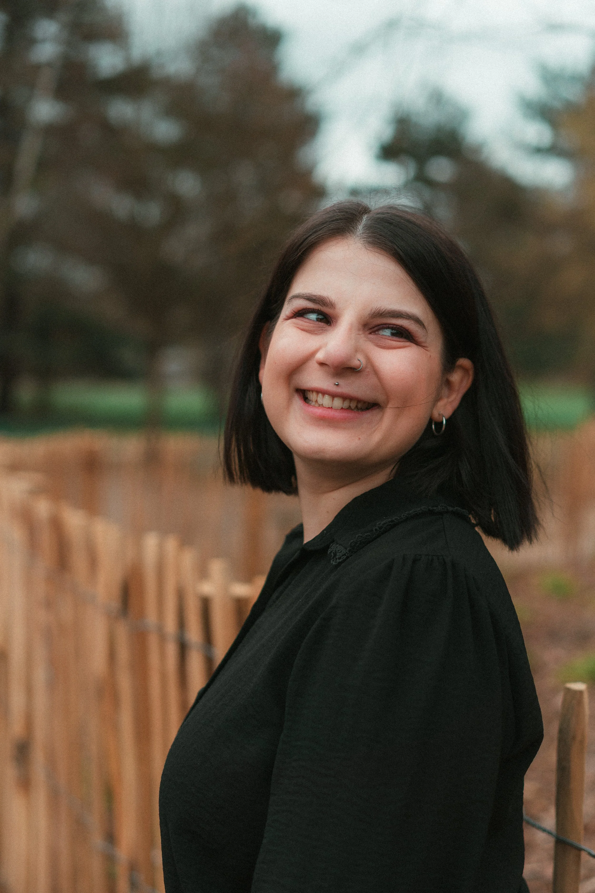Marlène photographe de mariage, portant une veste noire, se trouve à côté d'une clôture en bois dans un parc avec des arbres en arrière-plan.