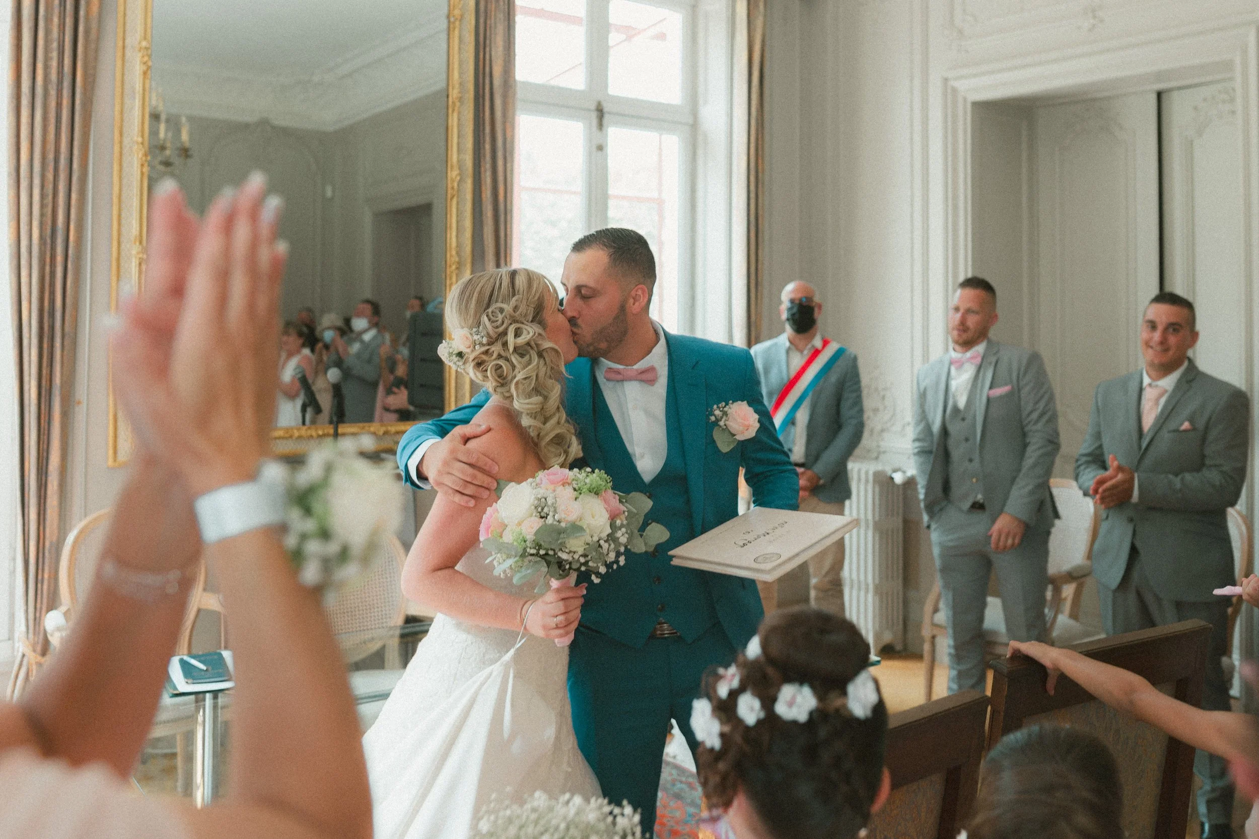 Un couple de mariés s'embrassent lors de leur mariage en intérieur, entourés d'amis et famille. La mariée porte une robe blanche et tient un bouquet, le marié porte un costume bleu avec un nœud papillon rose. Un grand miroir doré est en arrière-plan.