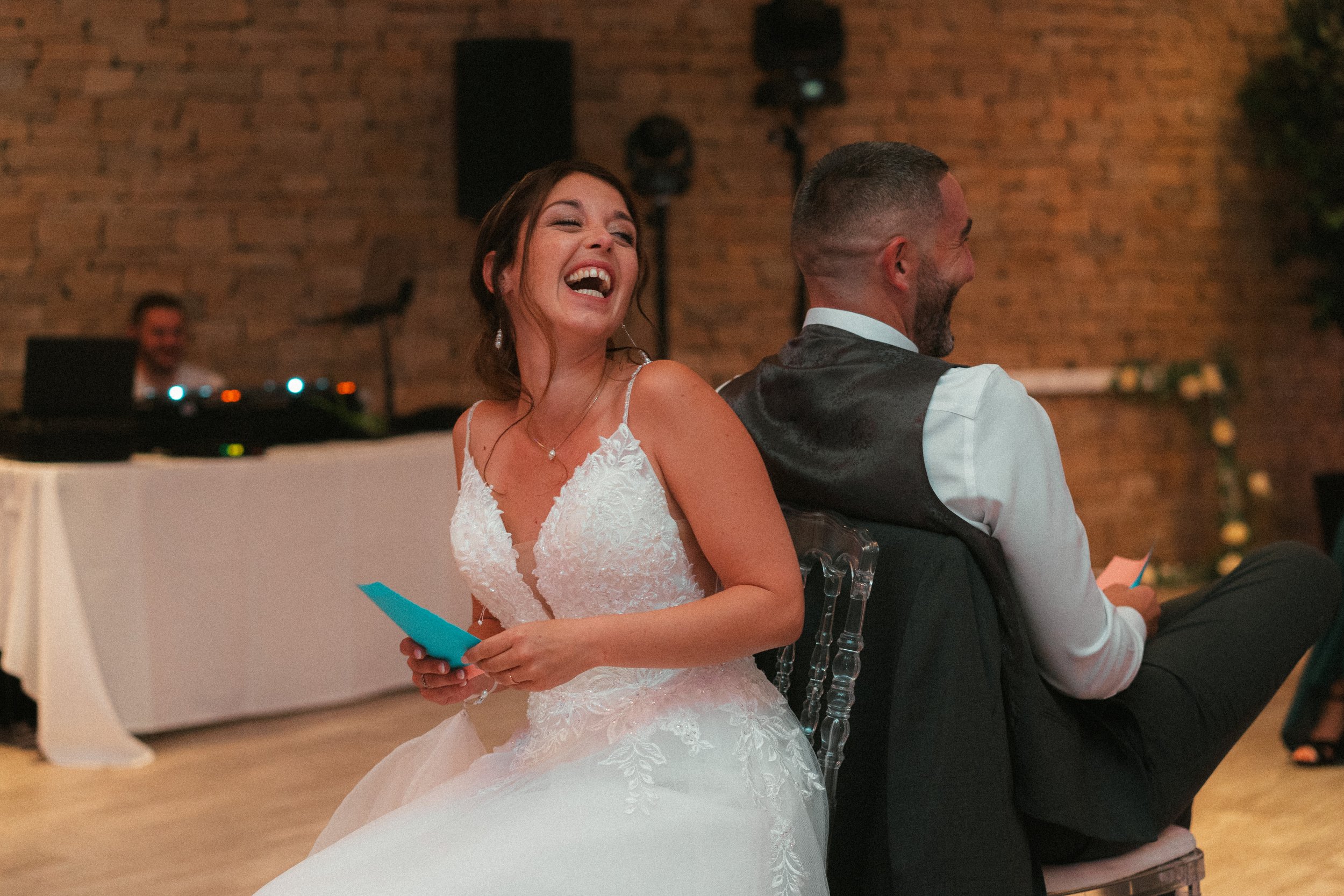 Une jeune femme en robe de mariée riant et souriant, assise à côté d'un homme en costume, lors d'une célébration de mariage à l'intérieur avec un mur en briques en arrière-plan.