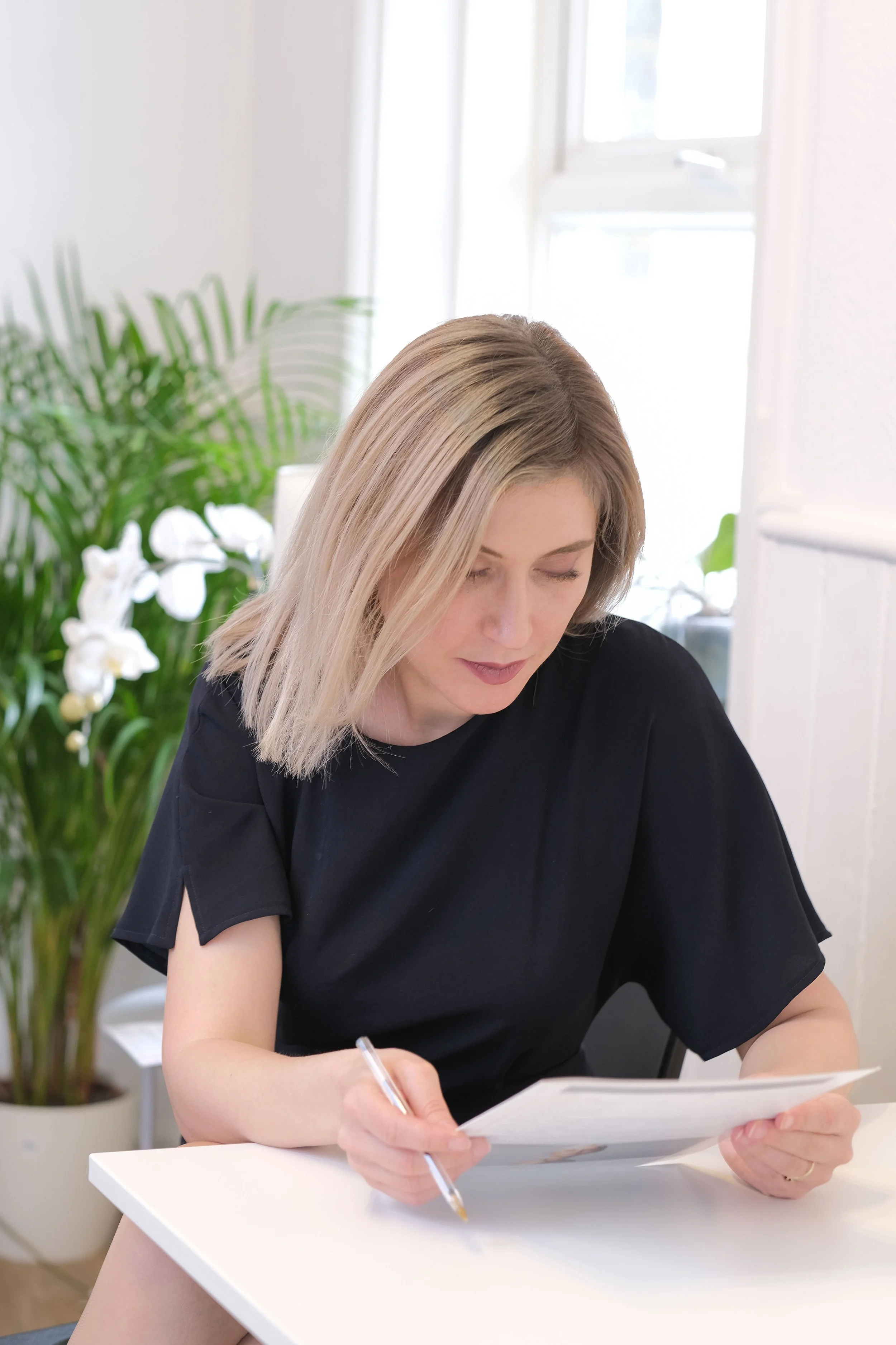 A woman with blonde hair sitting at a white table, reading a book, with a pen in hand, in a room with large windows and a potted plant with white flowers in the background.
