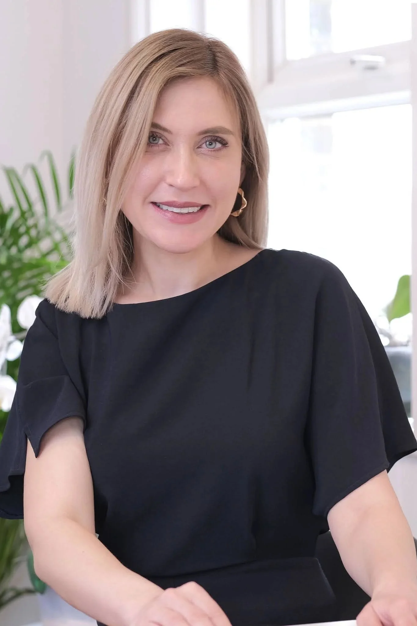 Close-up of a woman with shoulder-length blonde hair, wearing a black top, smiling, in a well-lit room with a window and green plants in the background.