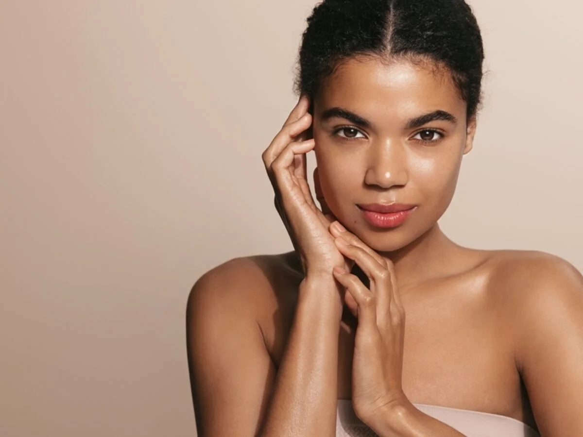 Close-up of a woman with natural hair and glowing skin, touching her face gently with both hands, looking at the camera against a beige background.