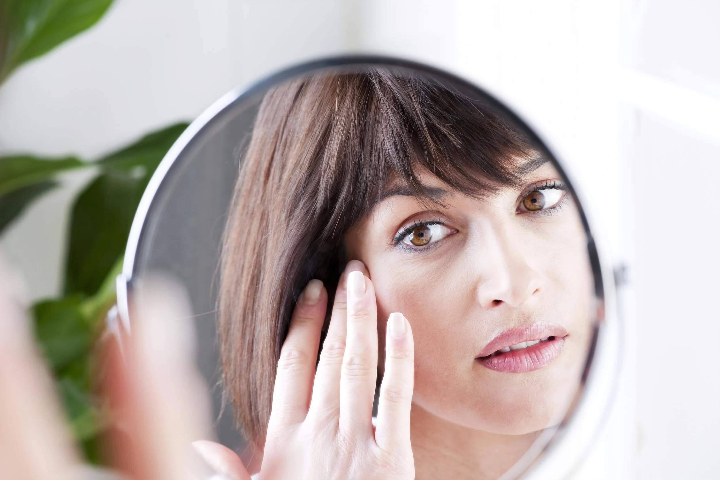 A woman with brown hair looking into a mirror while touching her cheek. There is a plant with green leaves partially visible on the left side of the image.
