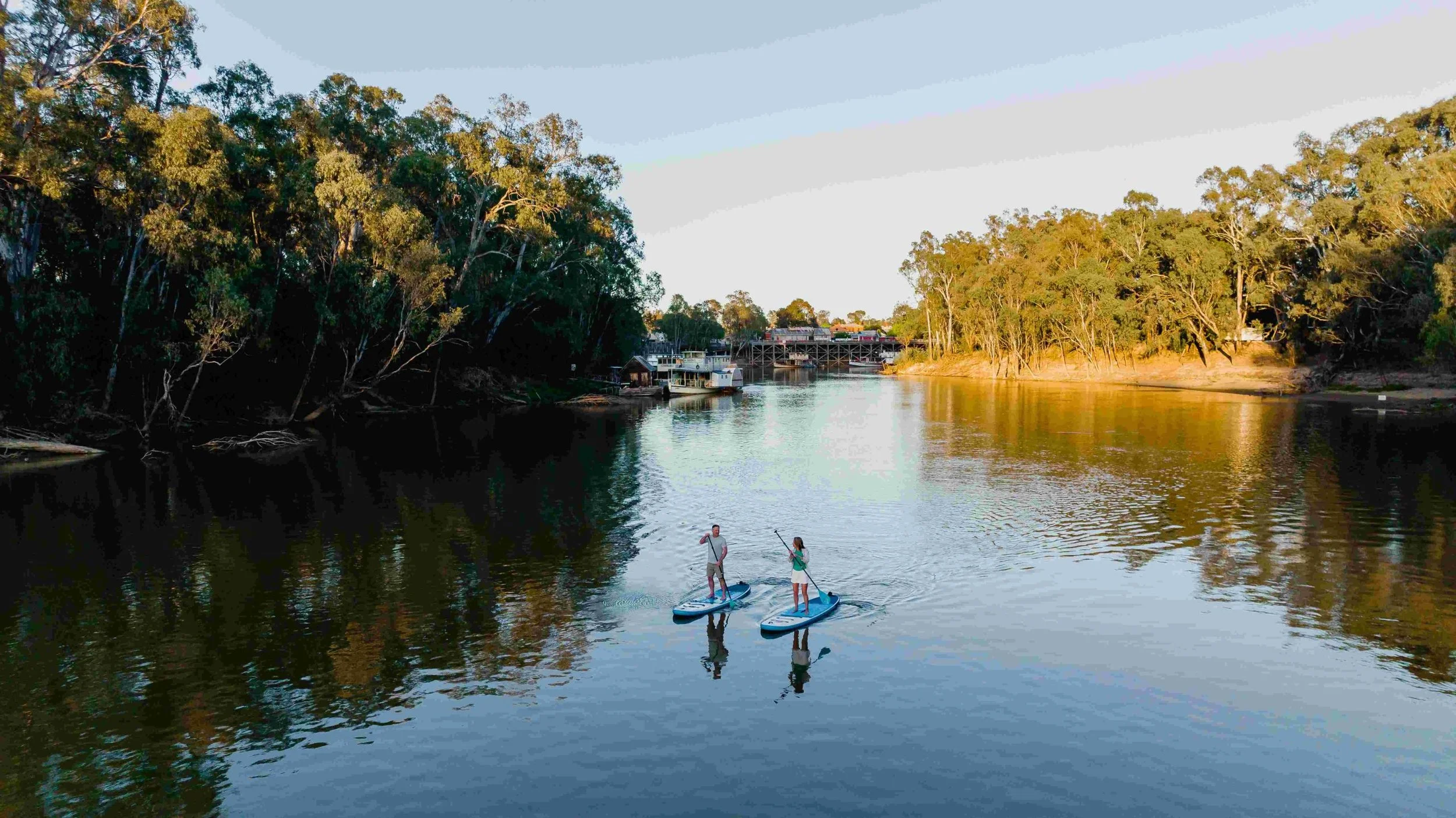 Two people stand on paddleboards on a calm river surrounded by trees, with houses visible in the background during sunset.