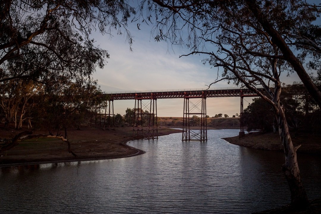 A red steel bridge over a calm river, surrounded by trees at sunset or dusk.