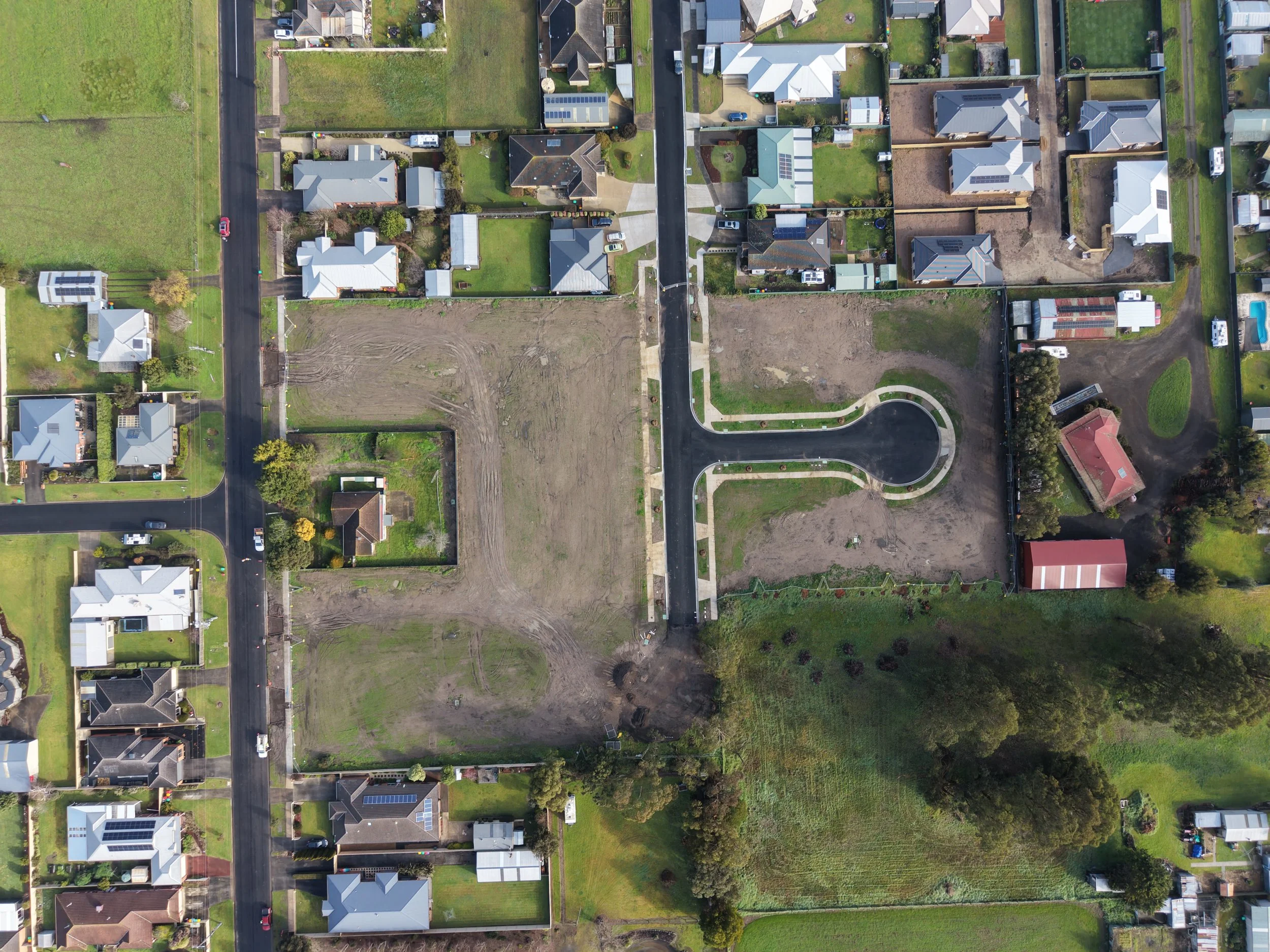 Aerial view of a suburban neighborhood with houses, yards, and streets. Some yards are lush and green, while others are under construction or vacant. Several houses have solar panels on the roofs. The streets include parked cars and sidewalks.