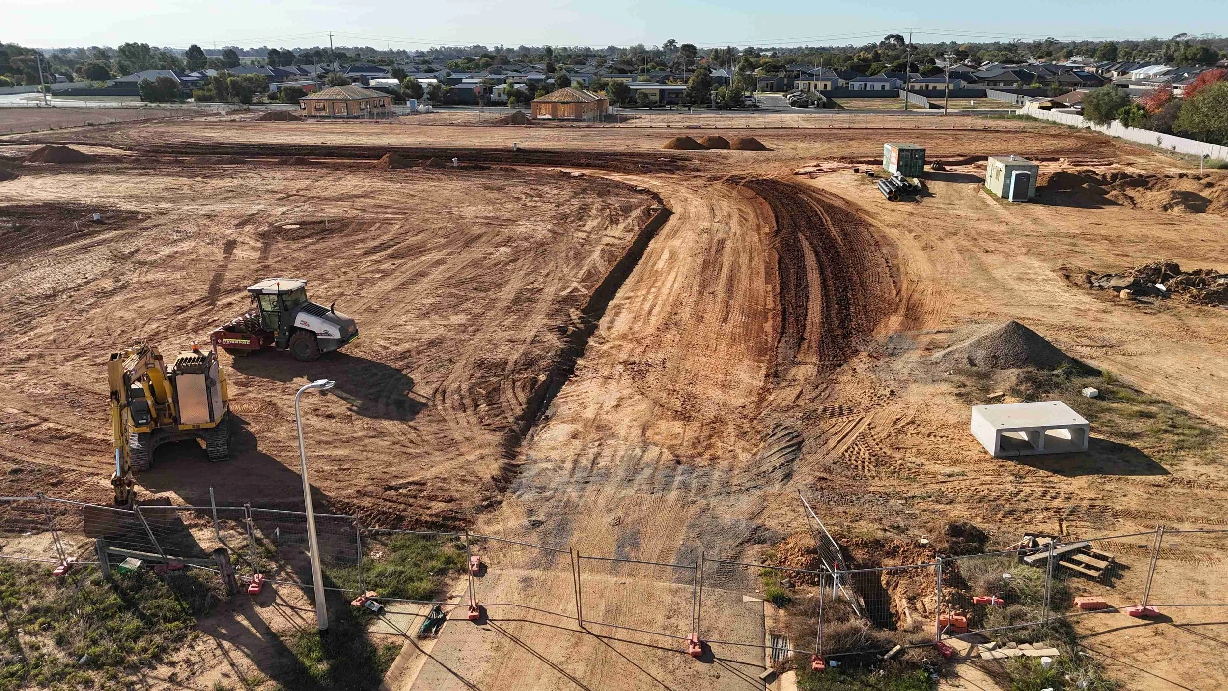 Construction site with heavy machinery and cleared earth, surrounded by a fence, near residential houses.