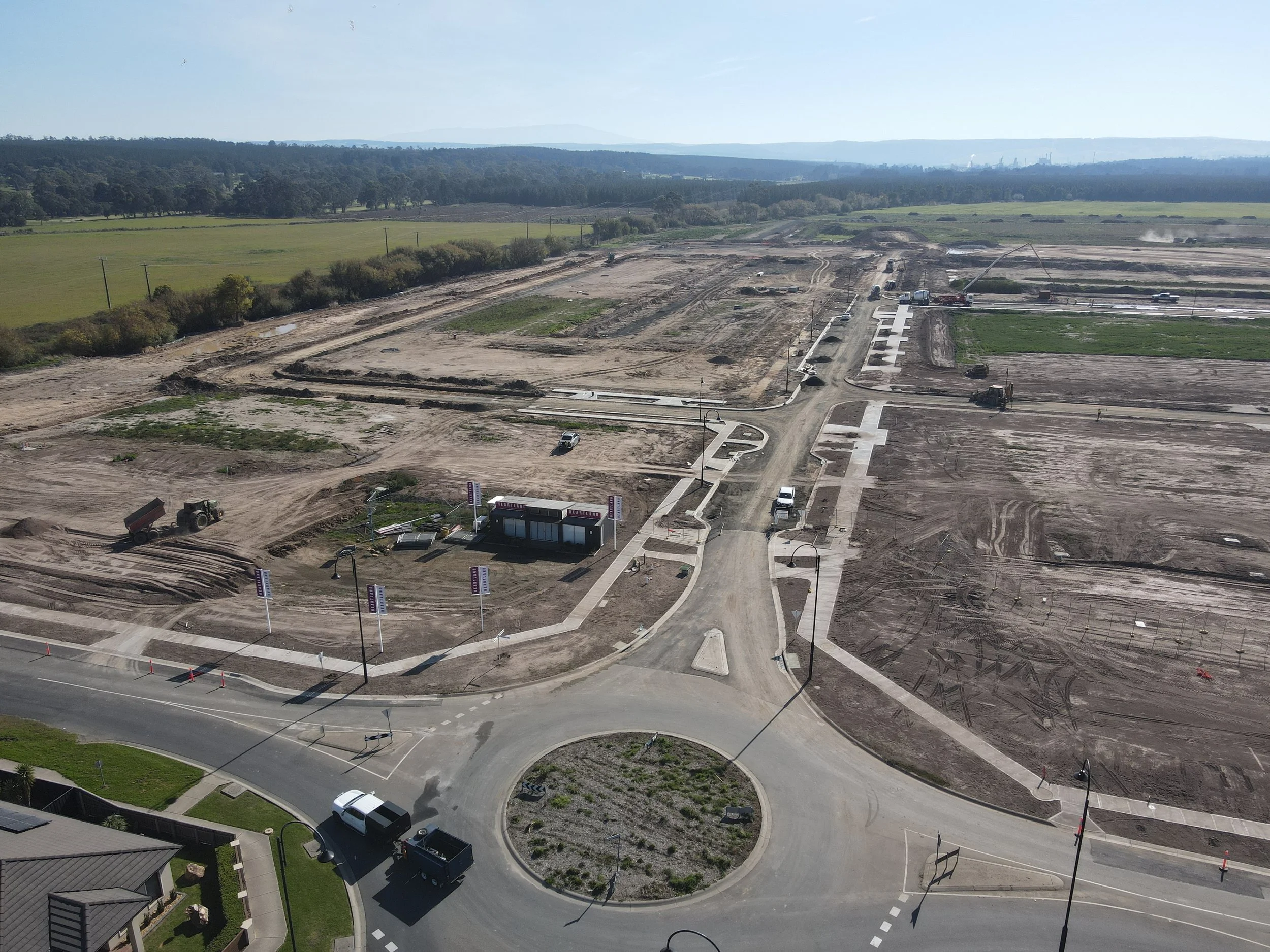 A construction site with cleared land, construction vehicles, and partially built roads, with a background of green fields and distant hills under a clear sky.