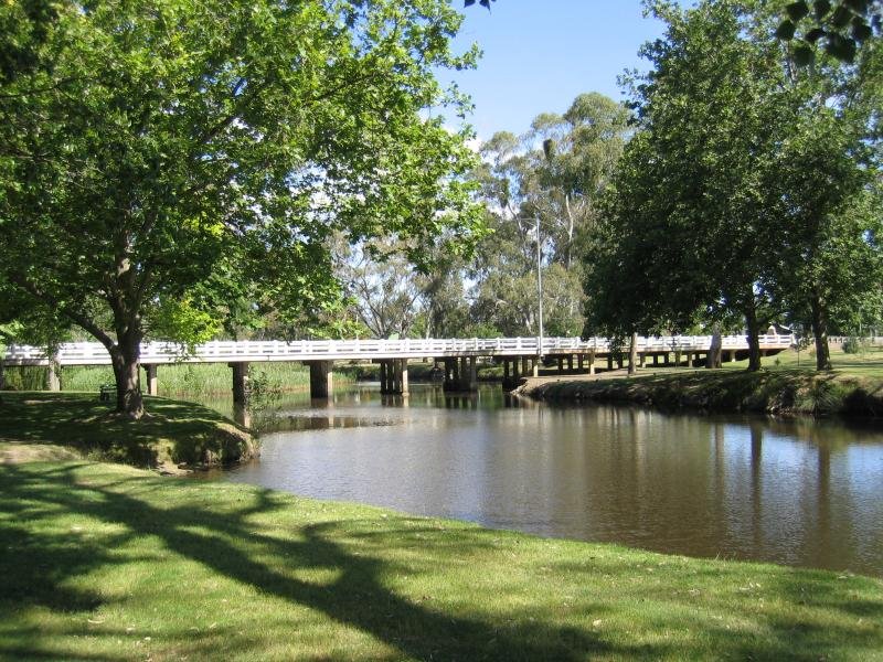 A peaceful park with green trees and grass, a pond, and a wooden bridge over the water.