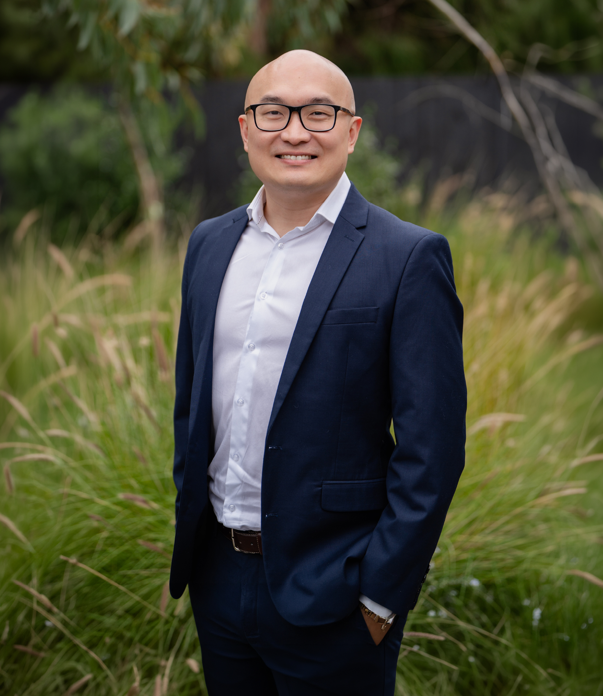Portrait of a man in a dark blue suit, white shirt, and glasses, smiling outdoors with green foliage and grass in the background.
