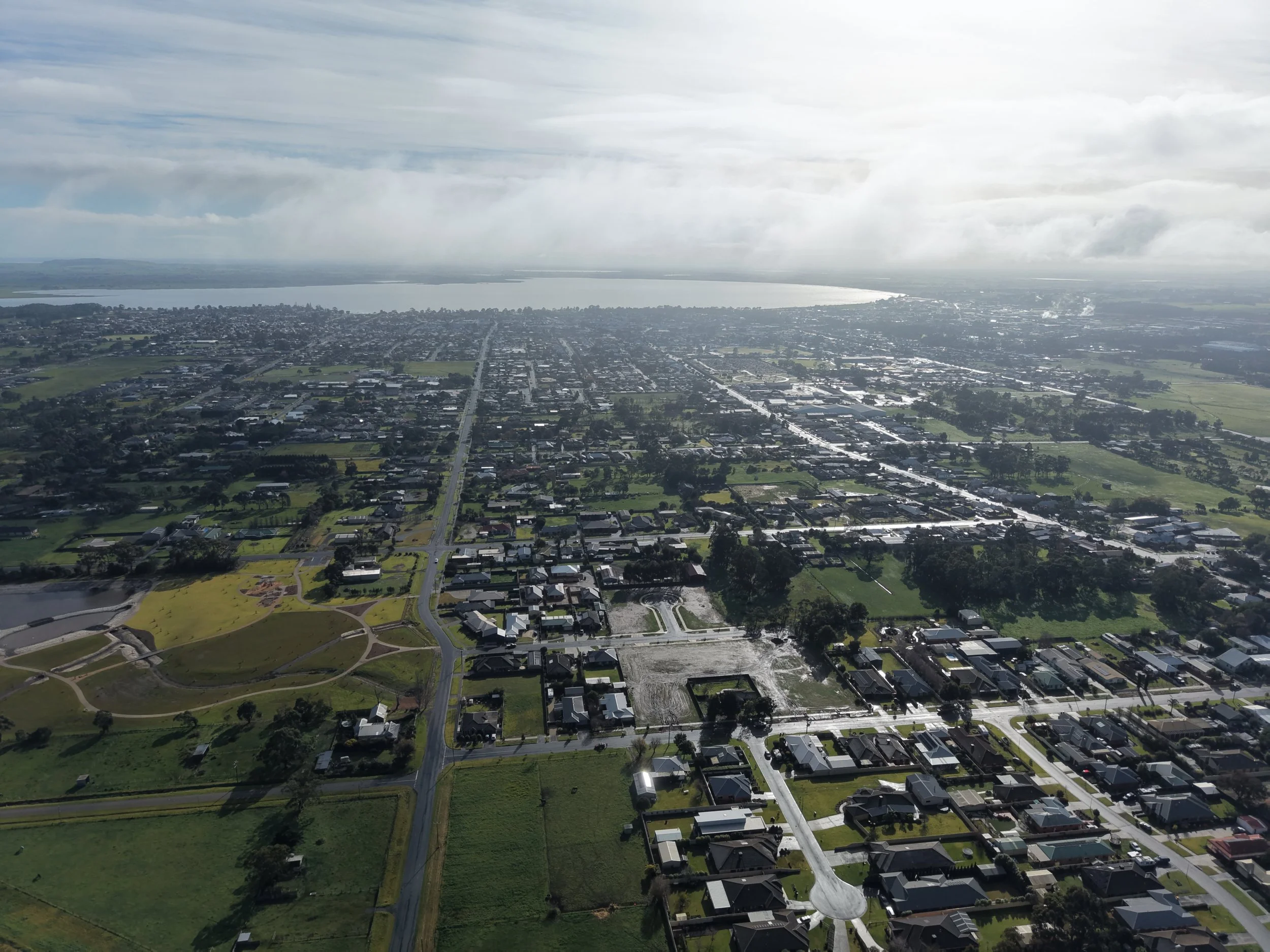 Aerial view of a suburban area with houses, roads, green parks, and a large body of water in the background, under a cloudy sky.