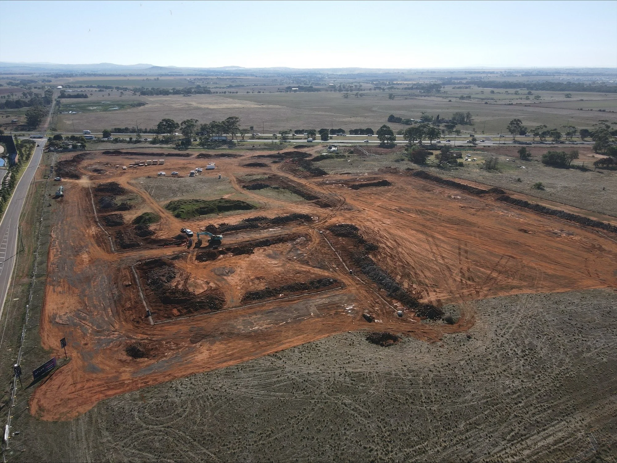Aerial view of a construction site with excavated areas and machinery, surrounded by open fields and a highway with vehicles.