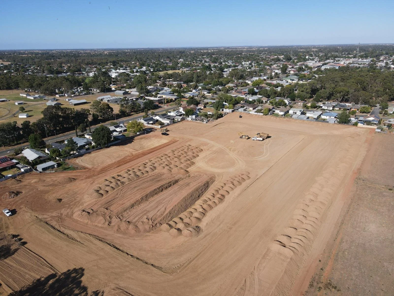 An aerial view of a construction site with dirt piles and construction vehicles, surrounded by a residential area with trees and houses.