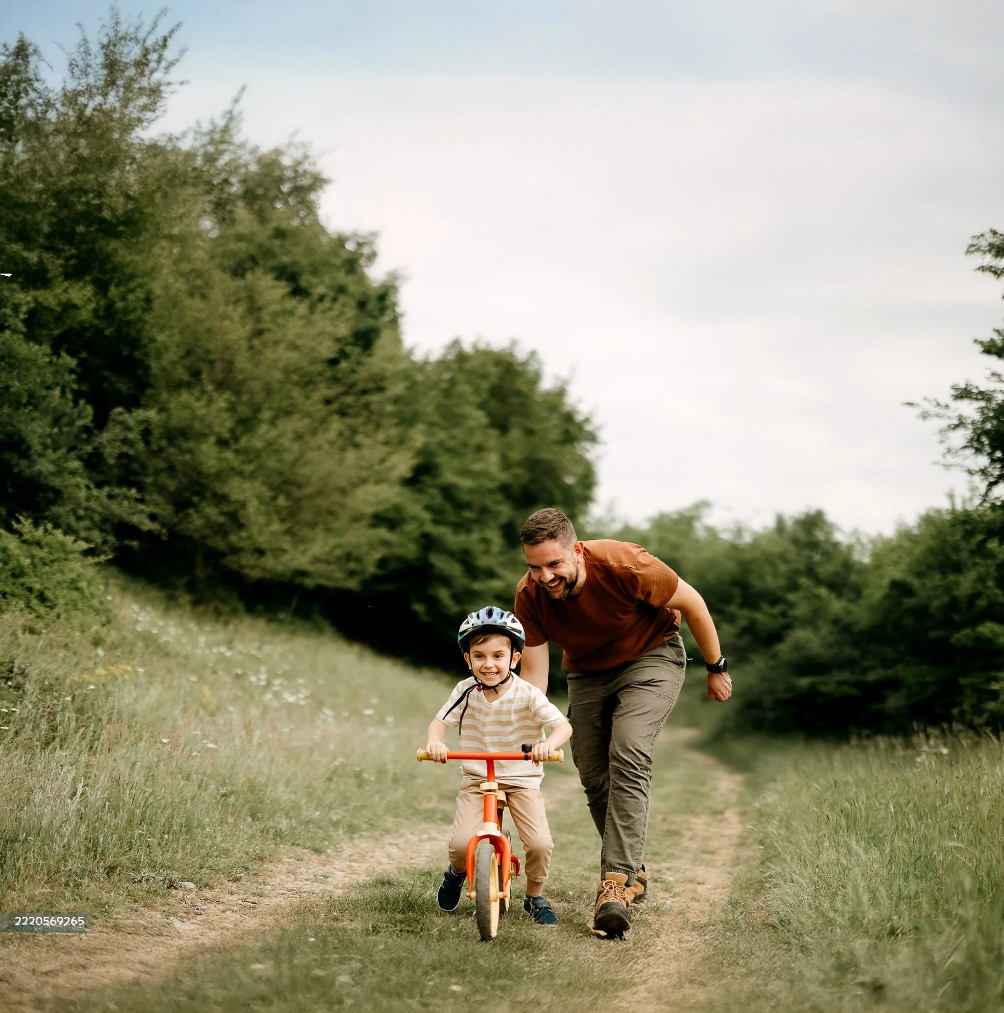 A man and a young boy riding a bicycle together on a dirt path surrounded by green trees, with the man smiling and helping the boy, who is wearing a helmet.