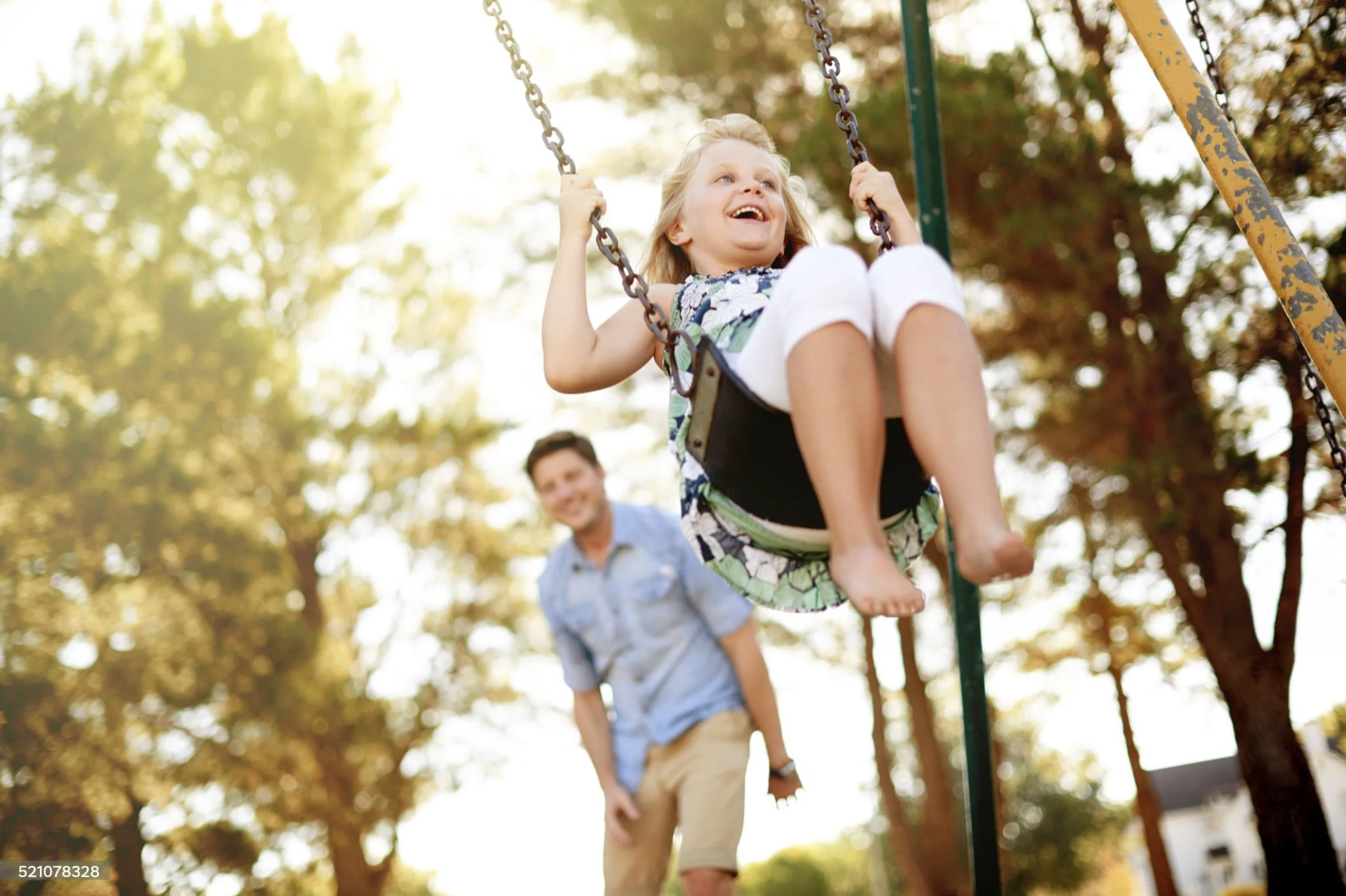 A young girl with blonde hair smiling as she swings on a playground swing, with a man behind her holding the swing. The scene takes place outdoors with trees and sunlight in the background.