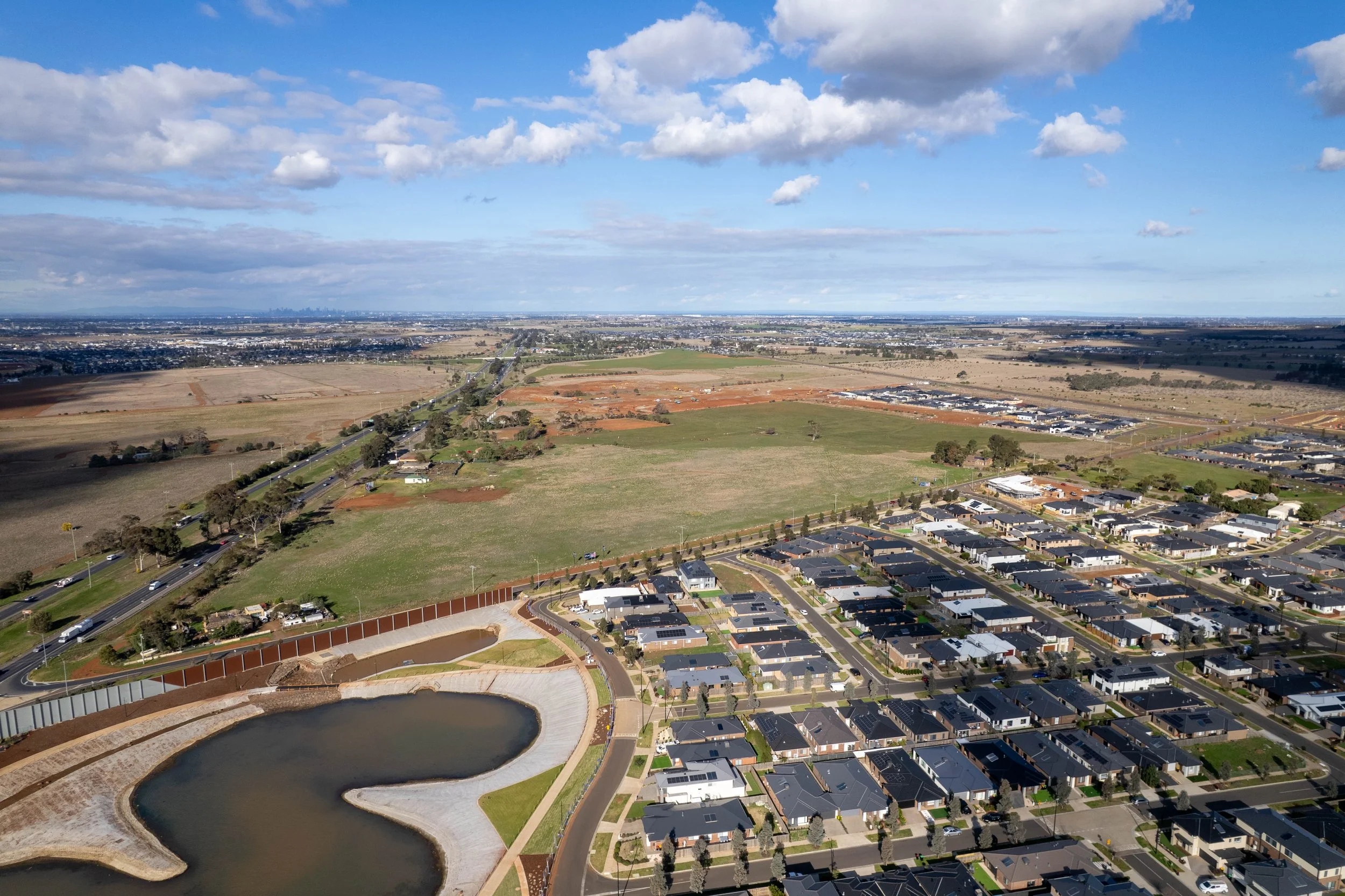 Aerial view of a suburban neighborhood with houses, a small lake, and open fields under a partly cloudy sky.