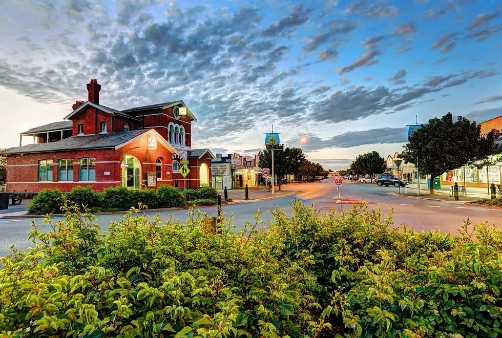 A small town main street scene at sunset with a red brick building on the left, surrounded by trees, bushes, and street signs, under a partly cloudy sky.