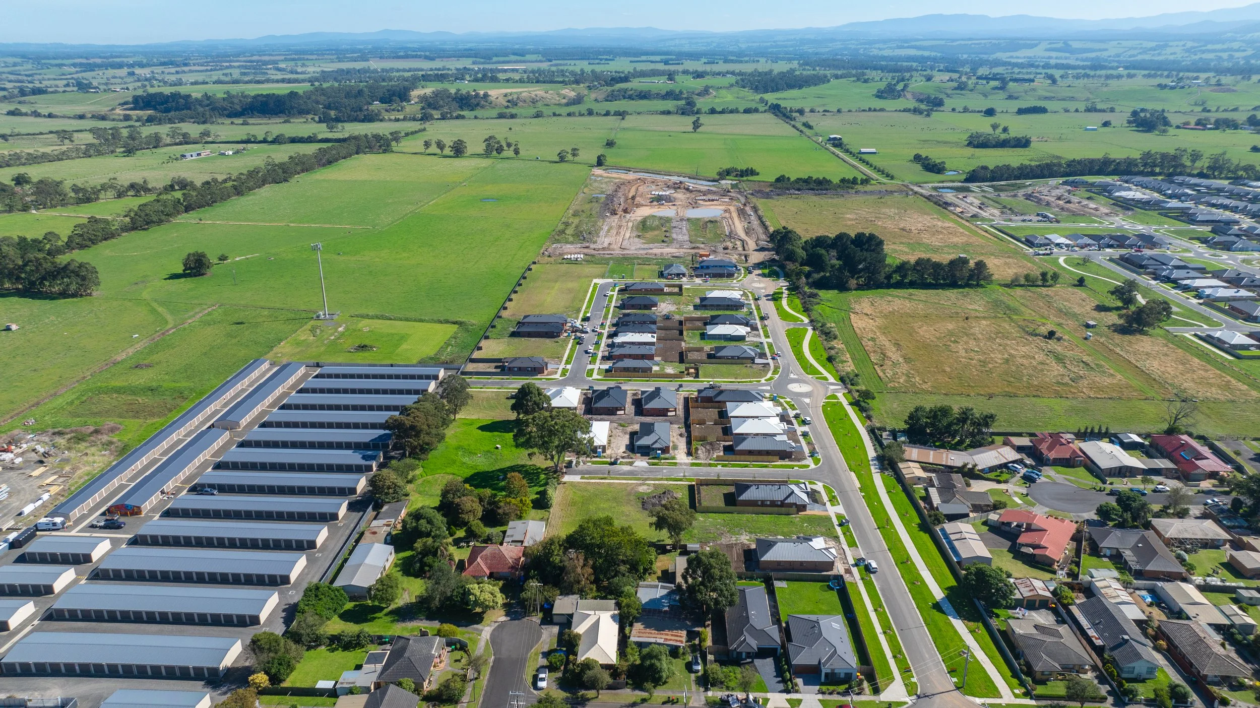 Aerial view of a suburban neighborhood with new residential houses, empty lots, and commercial buildings amidst green fields and farmland.