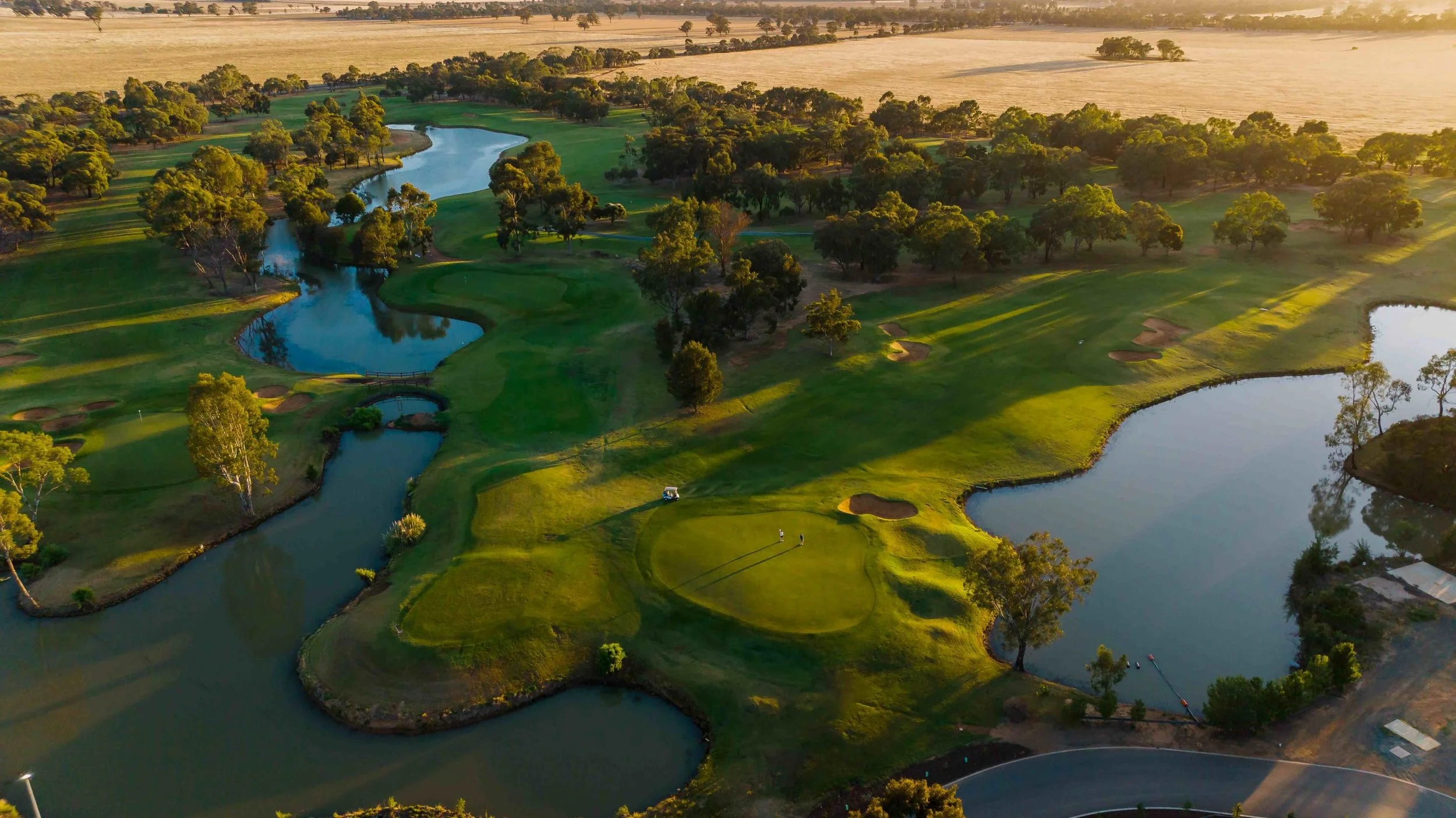 Aerial view of a golf course with water hazards, green fairways, sand bunkers, trees, and a golf cart on the course, surrounded by open fields.