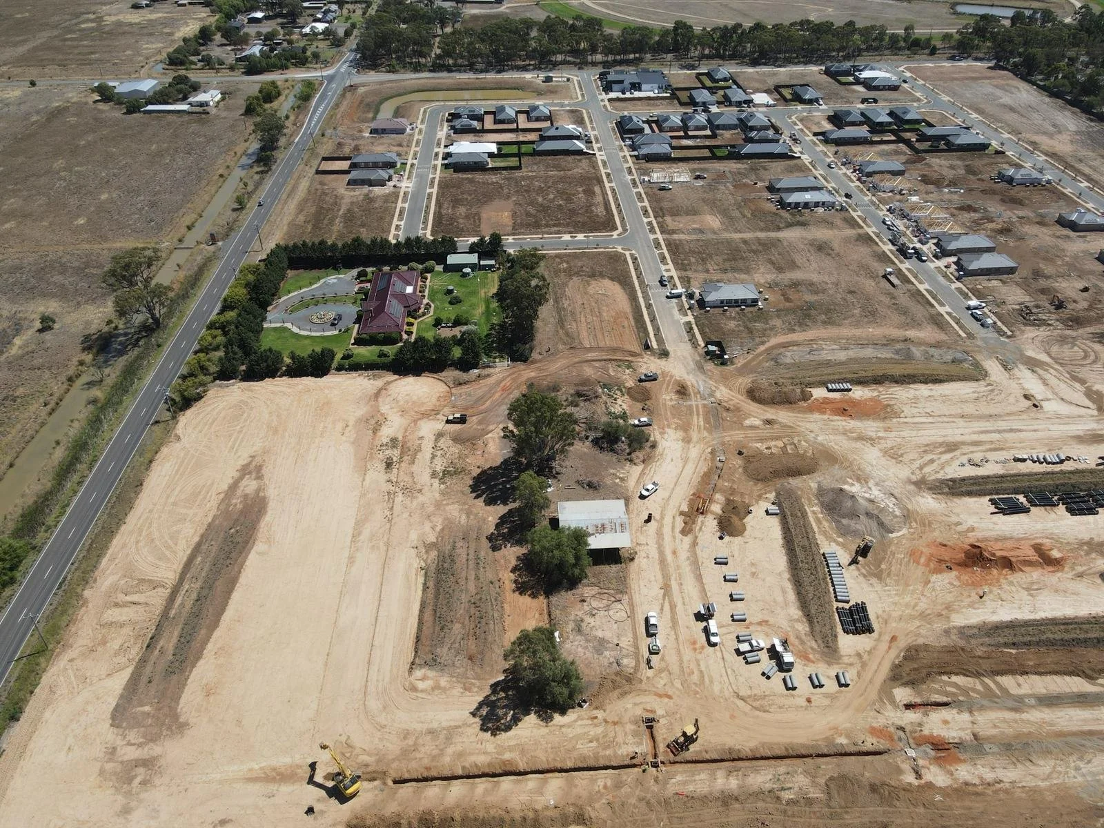 An aerial view of a construction site with cleared land and construction equipment, with a partially built residential area and a neighborhood with completed houses in the background.
