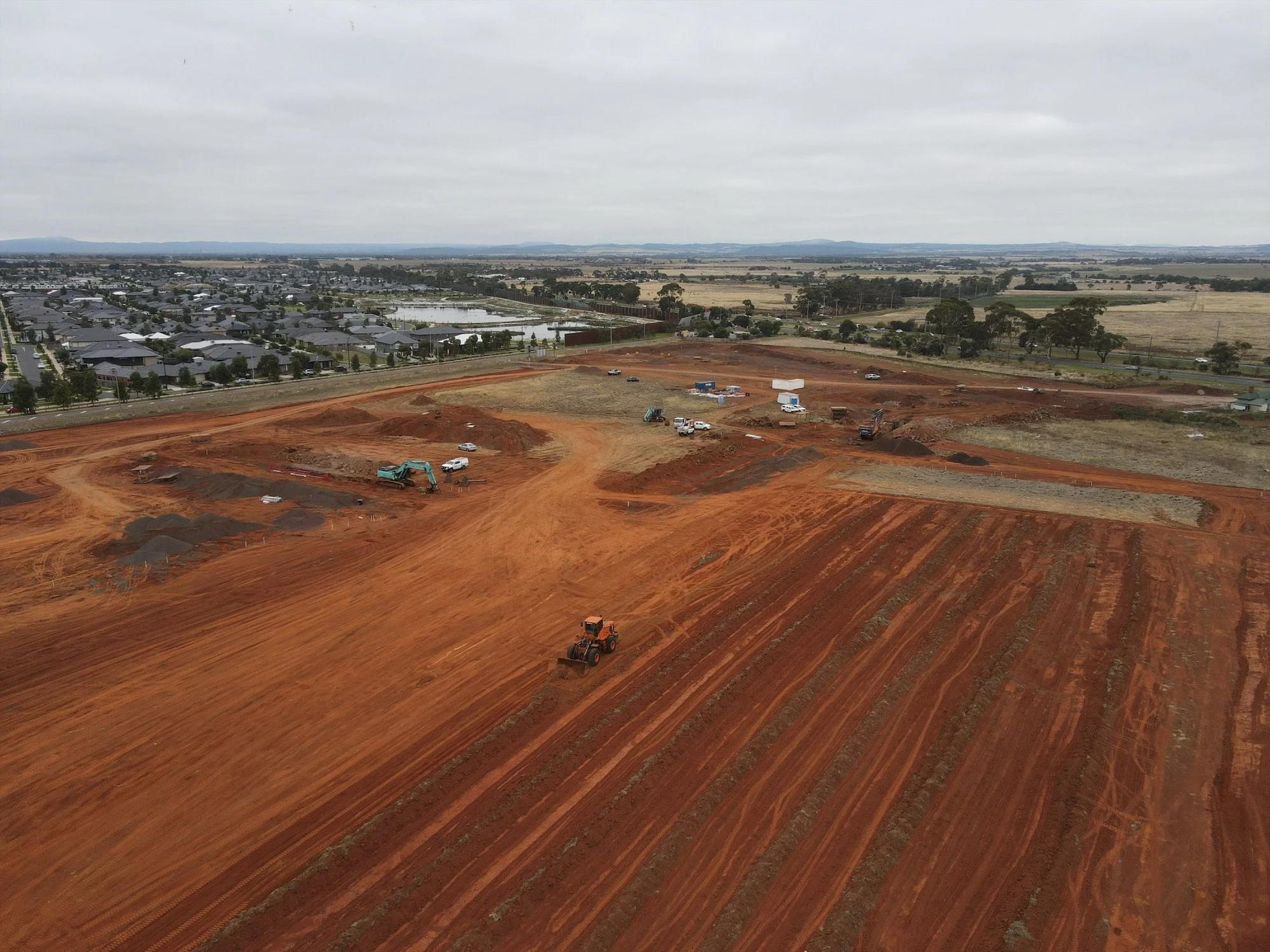 Construction site with red dirt and machinery, overlooking a suburban area with houses, a pond, and distant fields under cloudy skies.