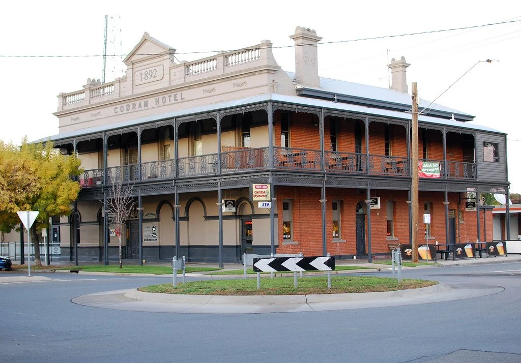 A historic two-story hotel with a sign reading 'Cobram Hotel' built in 1892, featuring a wraparound balcony with ornate ironwork, brick walls, and large windows, situated at a roundabout in a small town.