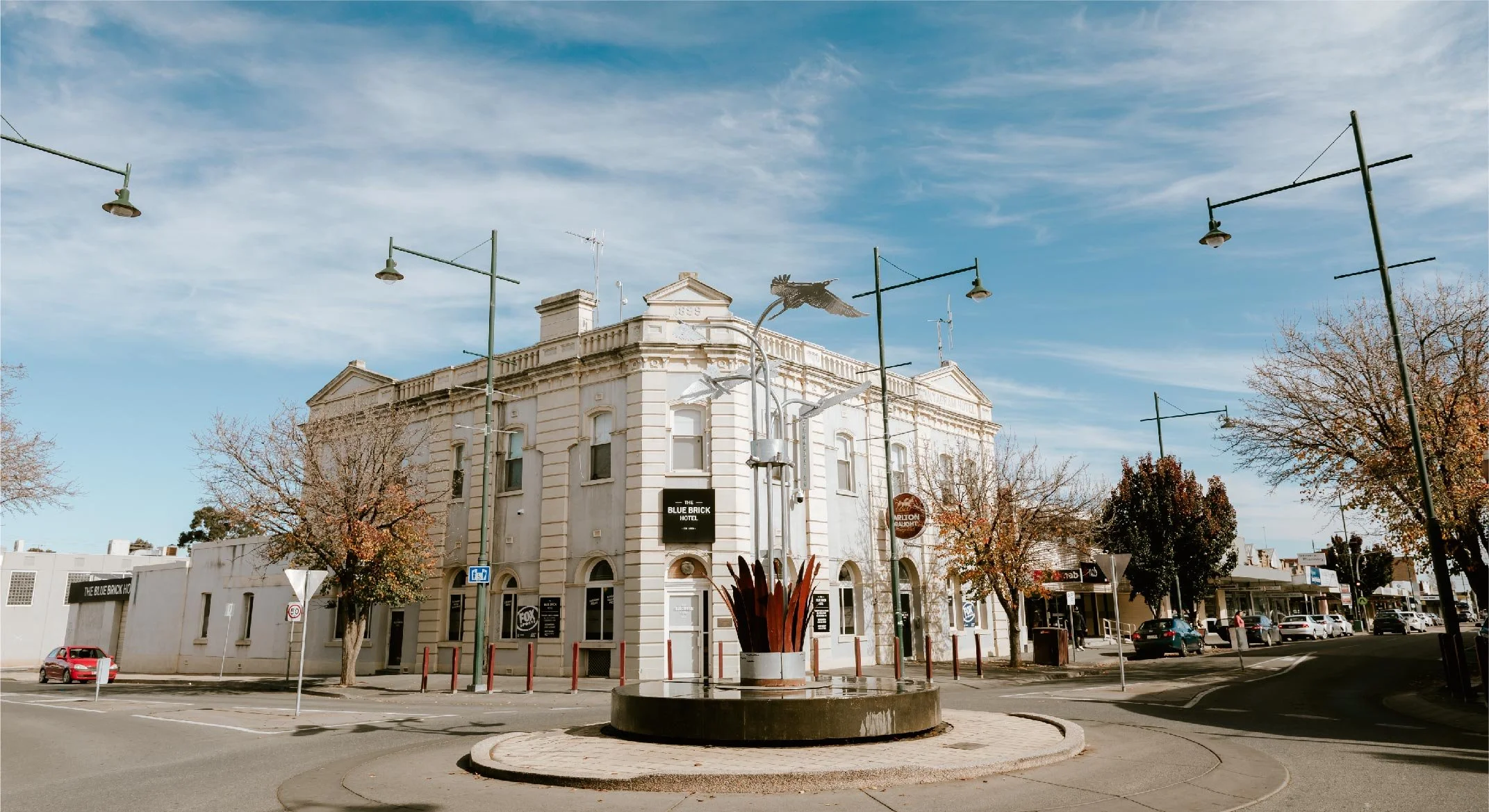 Historic white building with signs for the Blue Brick Hotel and Carlton Brewery, surrounded by trees with autumn leaves, street lamps, and parked cars under a clear blue sky.