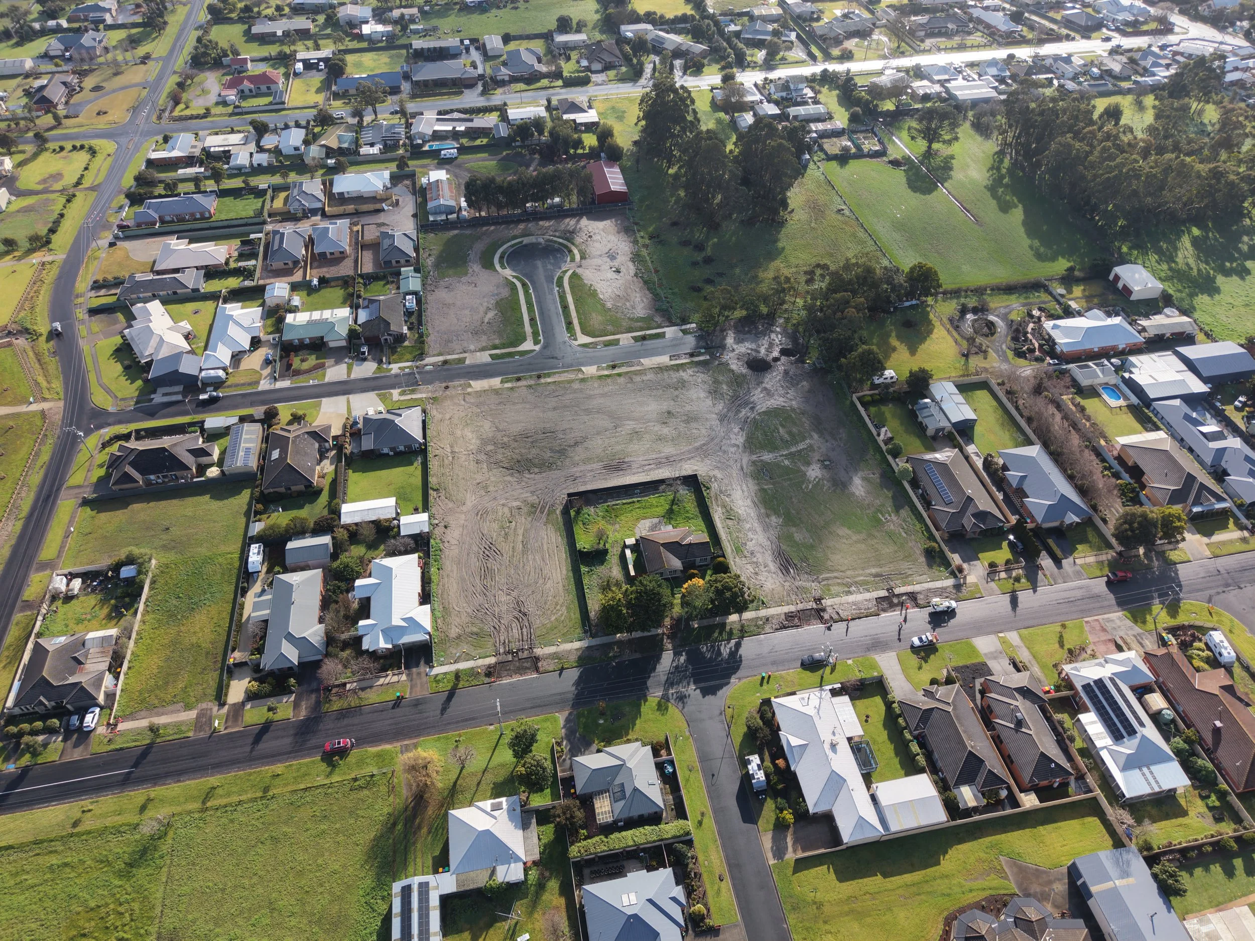 An aerial view of a suburban neighborhood with numerous houses, green lawns, and trees. Some houses have solar panels on the roofs. There are also undeveloped plots of land and a small roundabout. Streets and driveways connect the houses, with a few cars parked along the roads.