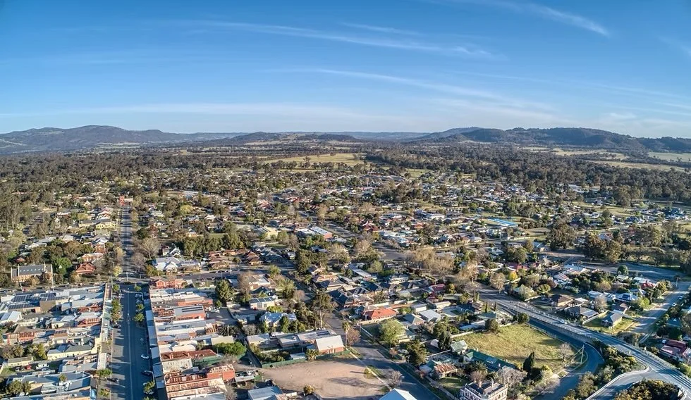 Aerial view of a suburban town with a mix of residential houses, streets, and green spaces, with hills in the distance under a partly cloudy sky.