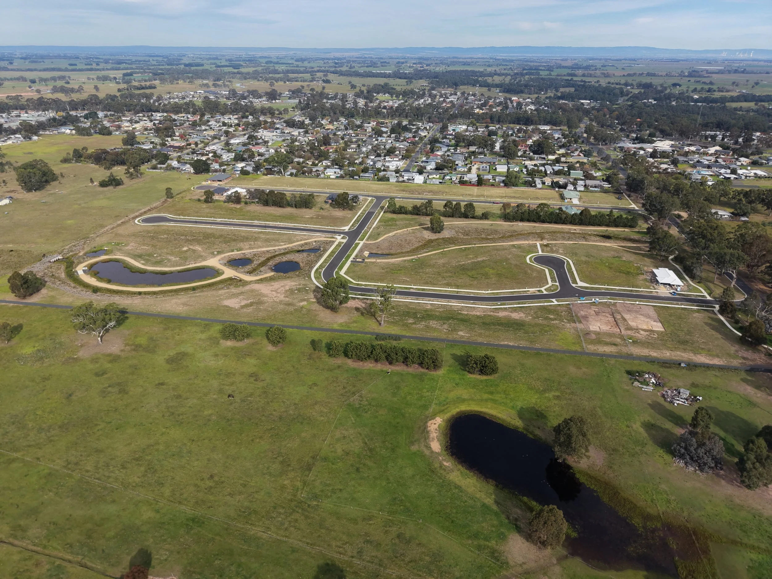 Aerial view of a small town with grid-like street layout, surrounded by green fields, a pond, and open land, under a partly cloudy sky.