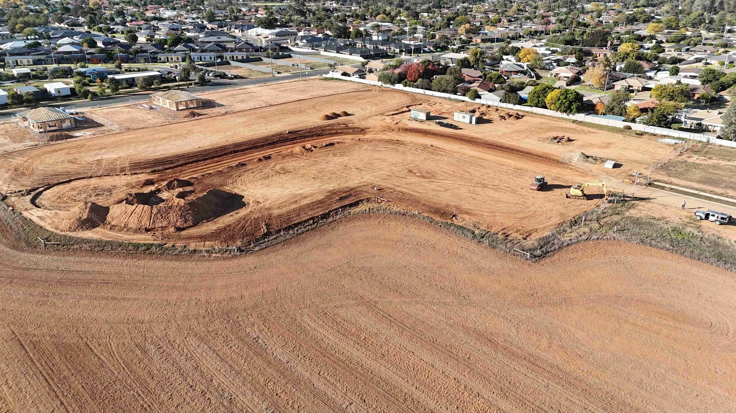 Aerial view of a construction site with cleared land, some small structures, construction vehicles, and dirt mounds, surrounded by a residential neighborhood with trees and houses.