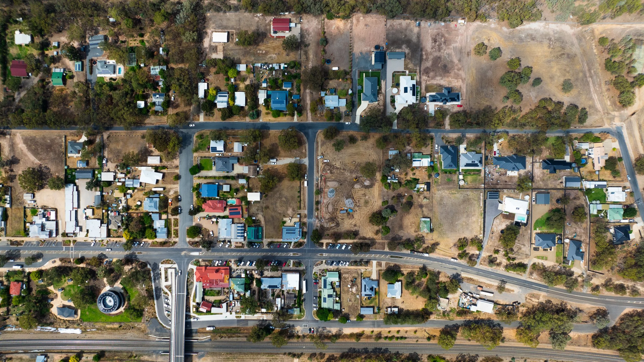 An aerial view of a neighborhood with various houses, trees, and streets, with a highway running at the bottom of the image.