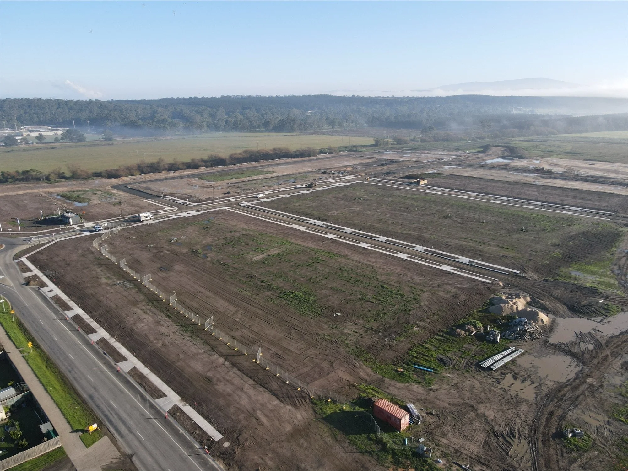 An aerial view of a construction site with cleared land, construction materials, and roadways, with hills and forests in the background.