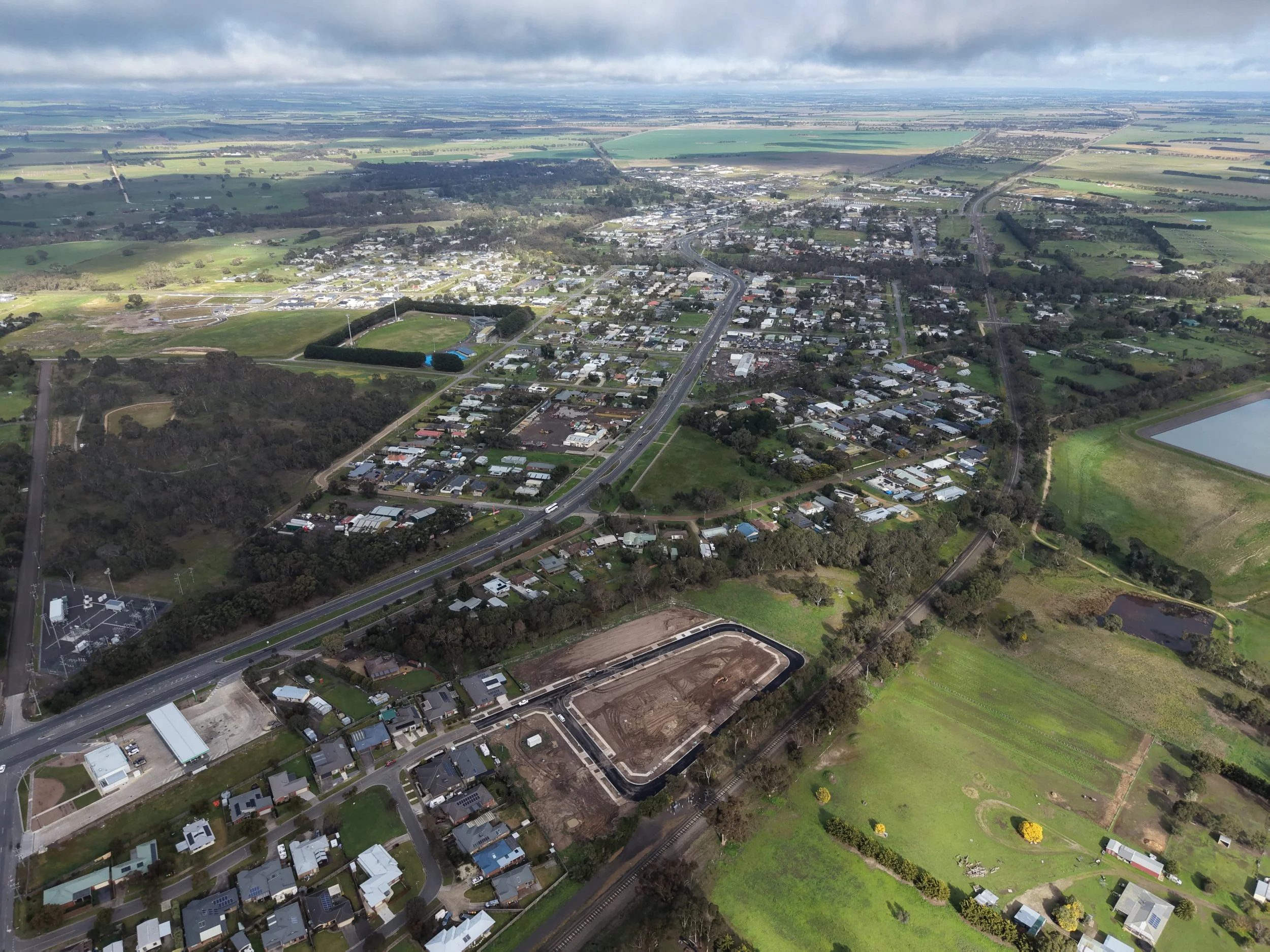 Aerial view of a small town with residential and commercial buildings, roads, green parks, and surrounding fields under a partly cloudy sky.