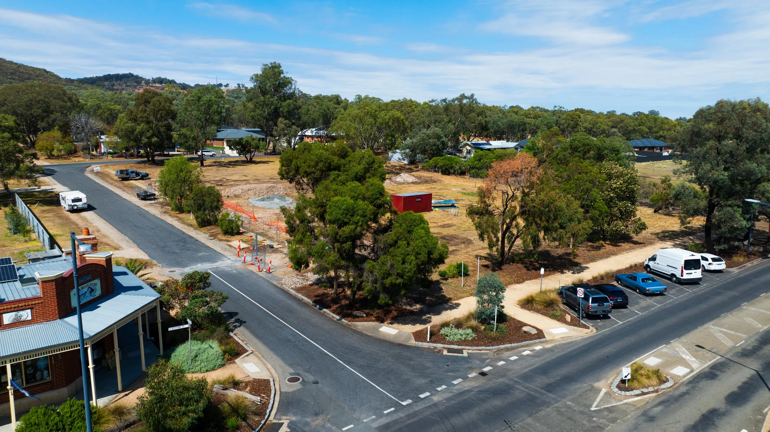 Aerial view of a parking lot and street in a suburban area with trees and houses, some construction work with orange barriers and equipment visible.