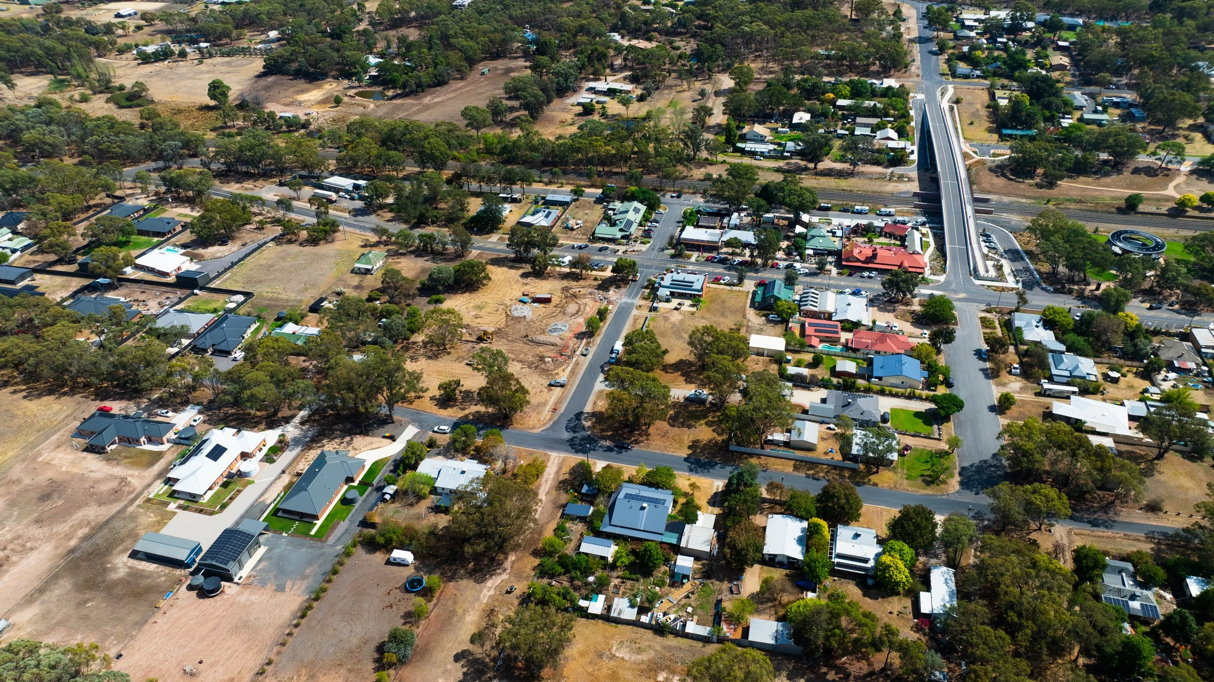 Aerial view of a residential neighborhood with houses, trees, and roads, including a bridge over a railway track and a roundabout in the top right corner.