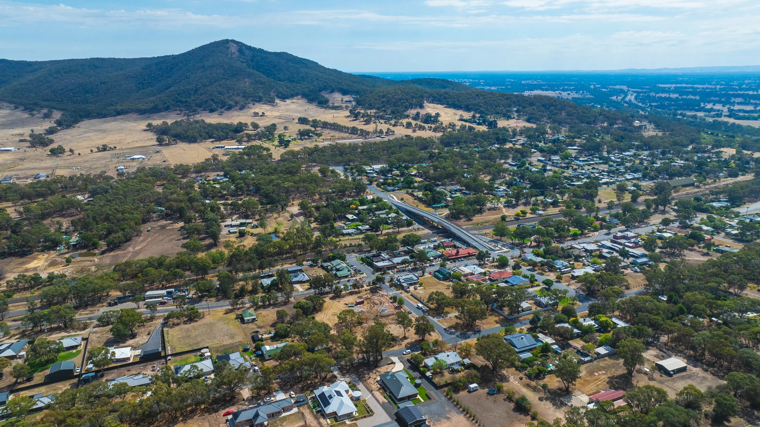 Aerial view of a small town with houses, trees, and roads, surrounded by hills and a mountain in the background.