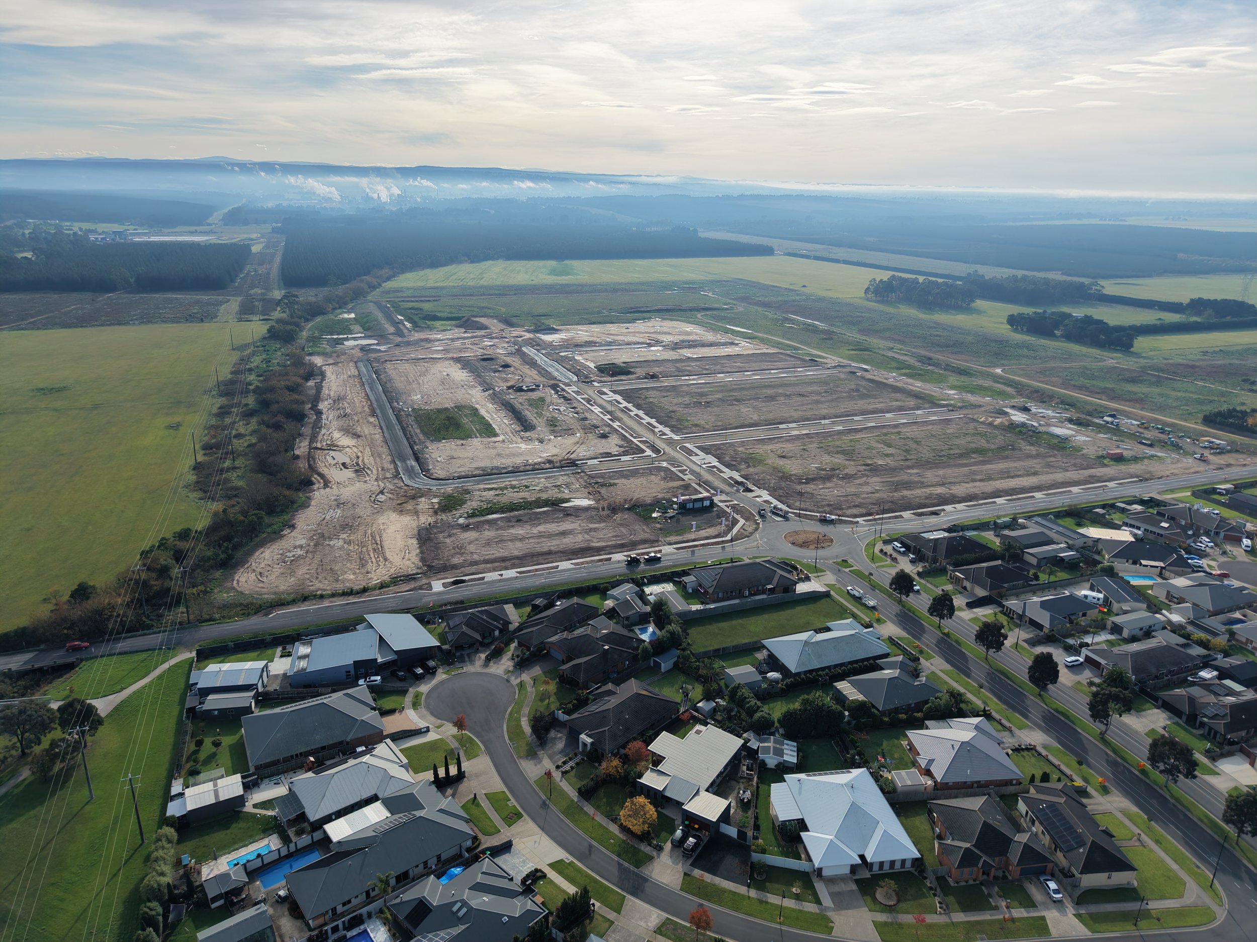 An aerial view of a suburban neighborhood with houses, some with pools, and a construction site in the background. The area is surrounded by fields, trees, and distant industrial smoke plumes under a partly cloudy sky.