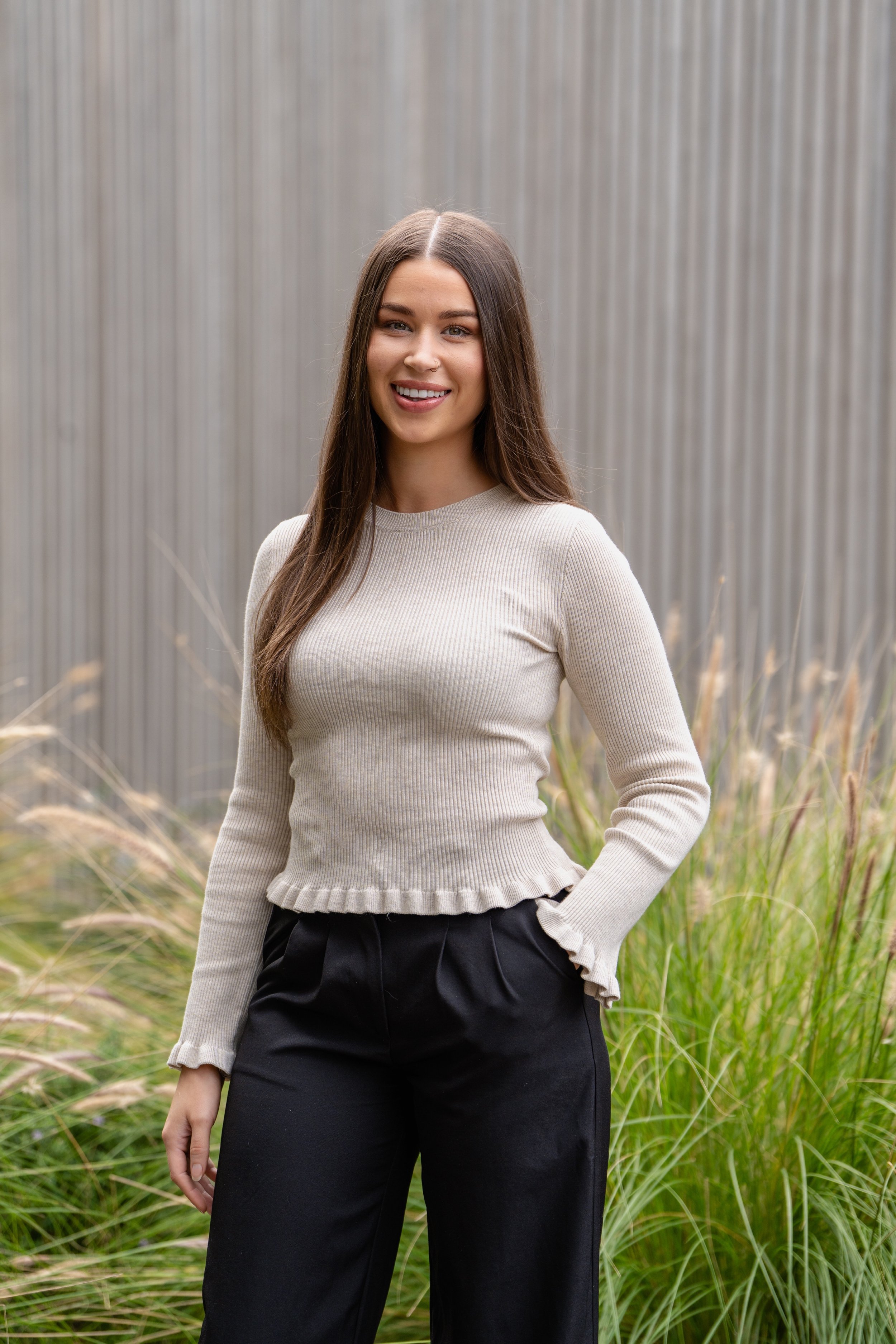 A young woman with long brown hair, wearing a beige long-sleeve top and black pants, standing outdoors in front of a metal wall and tall grass, smiling at the camera.