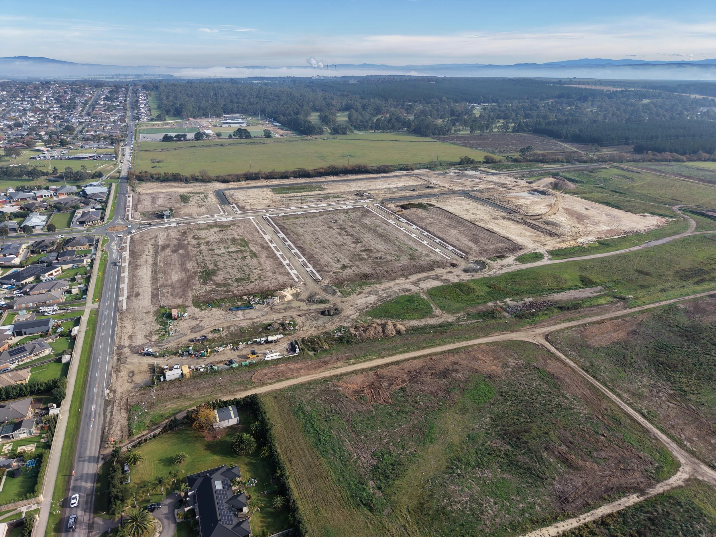 Aerial view of a developing construction site with cleared land and ongoing groundwork, neighboring a suburban residential area with houses, roads, and green fields, with a backdrop of forest and distant mountains.