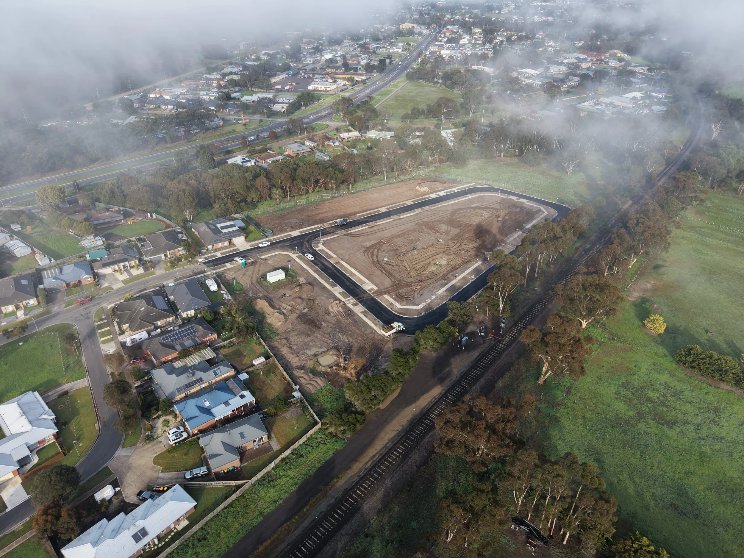 Aerial view of a residential neighborhood with houses, trees, and a foggy atmosphere. There is a construction site with dirt and paving in progress, and a railway track runs alongside the area.