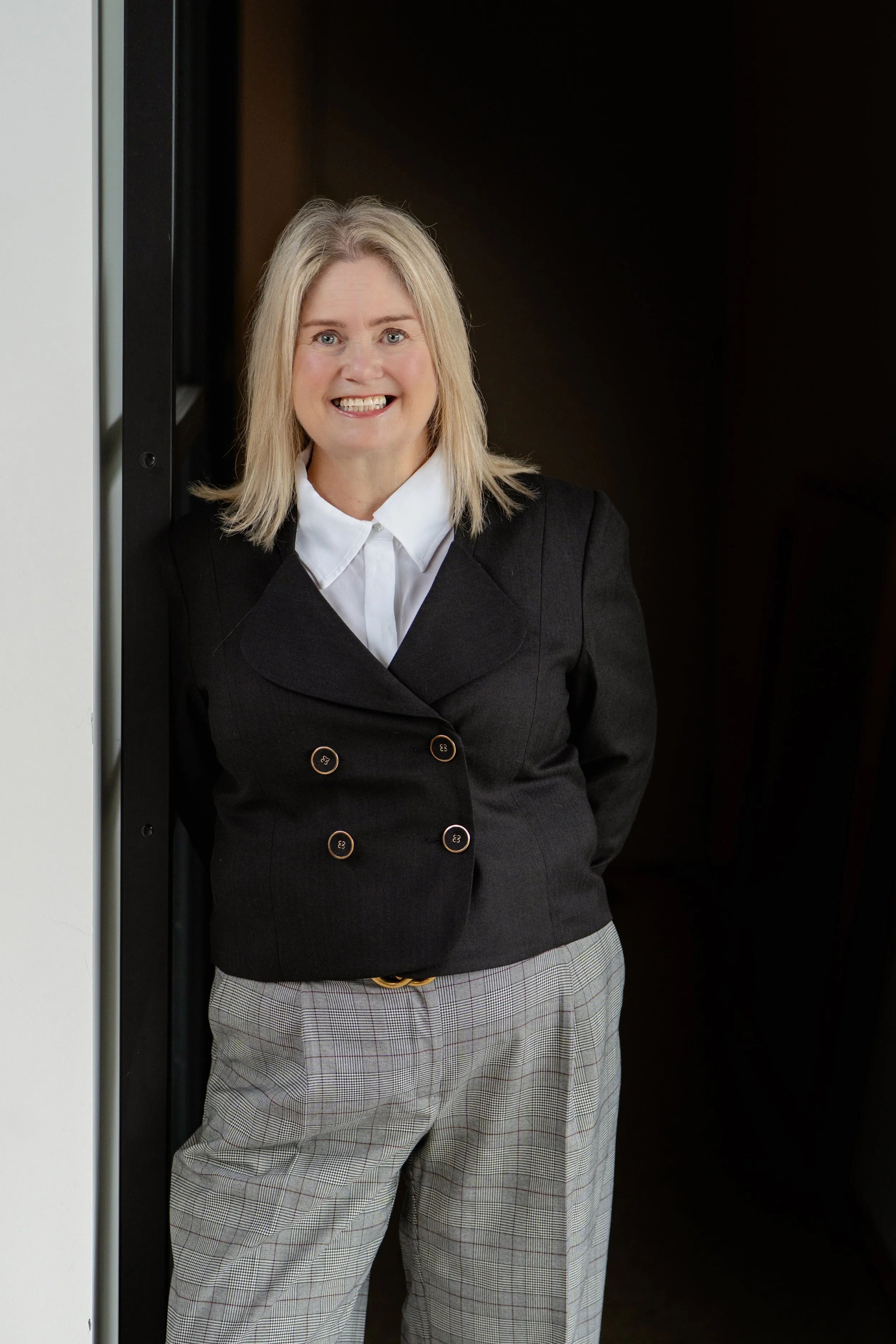 A woman with blonde hair smiling, standing in an elevator doorway. She is wearing a black blazer with gold buttons, a white shirt, and plaid pants.